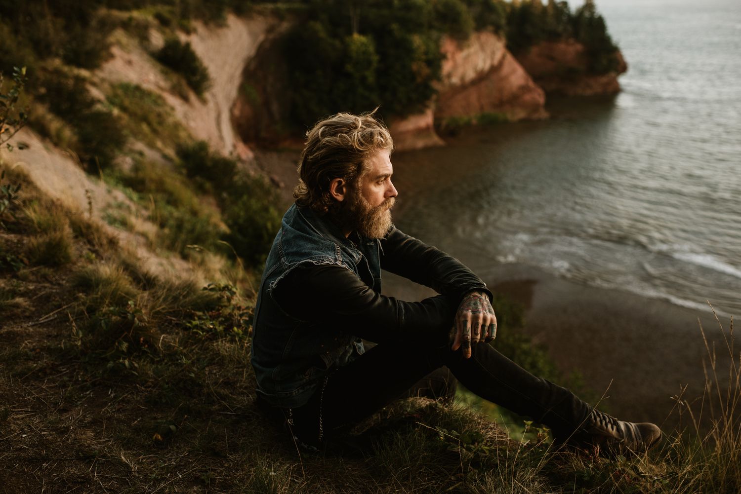 Bearded man sitting on coastal cliffside grass, gazing at ocean with red rocky cliffs in background.