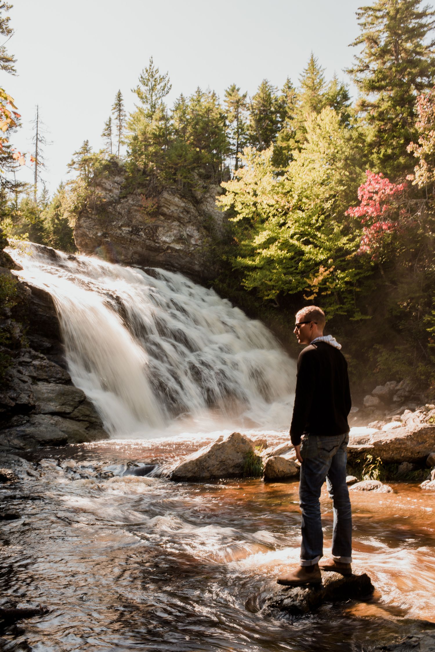 Man standing on rocks admiring a scenic waterfall surrounded by autumn trees in a forest.