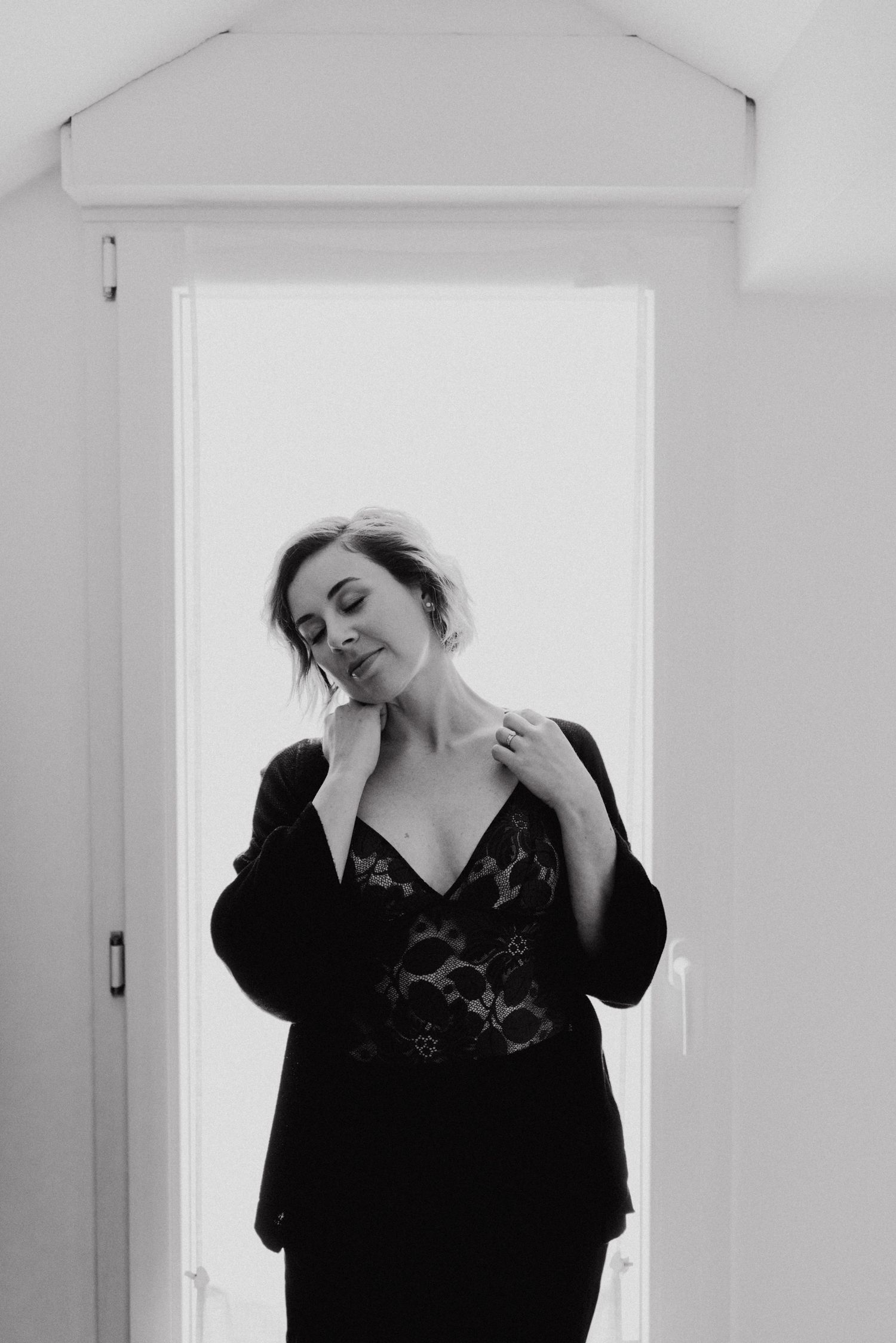 Black and white photo of a woman in a lace top and blazer standing in a doorway with soft natural light.