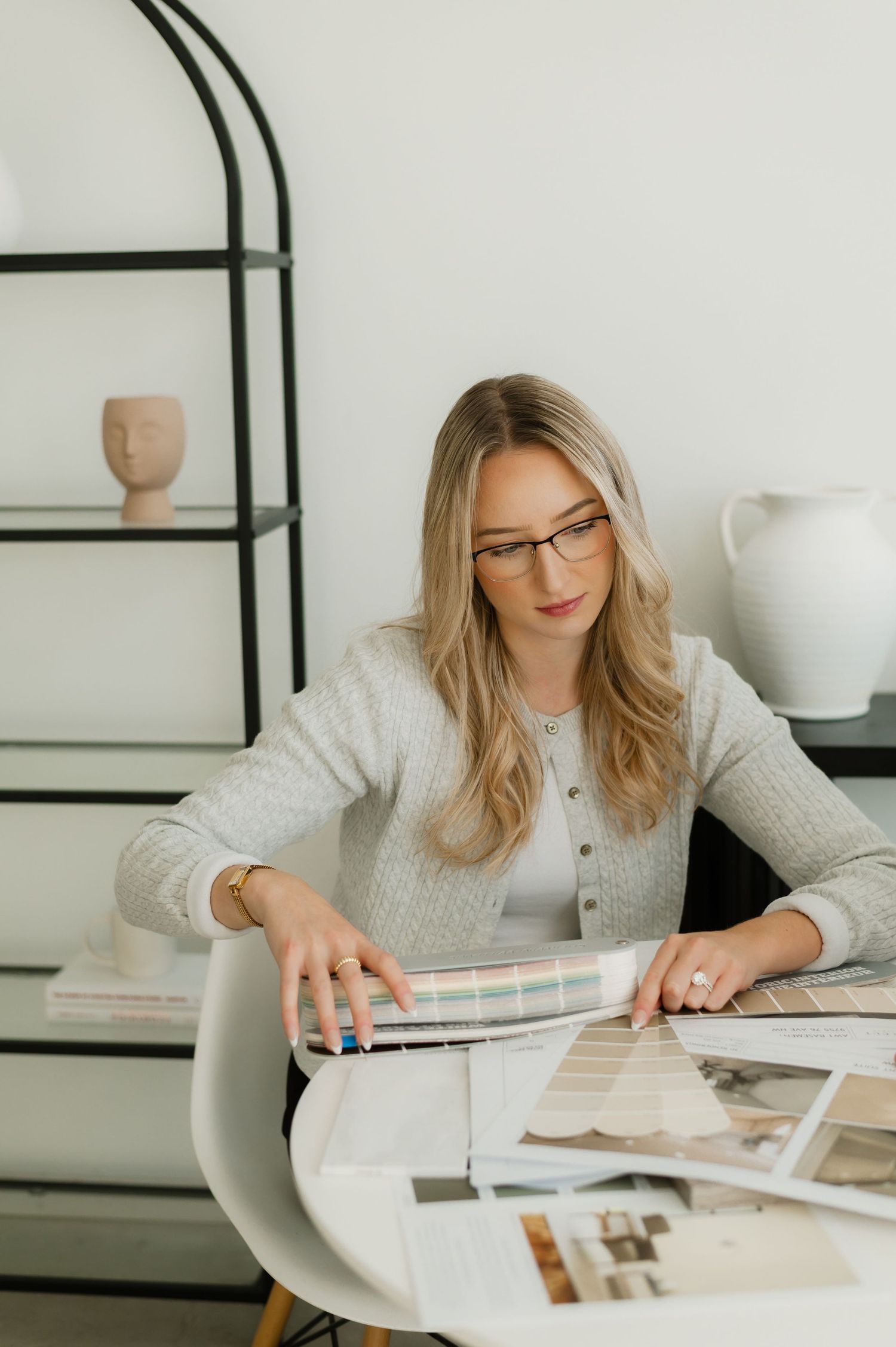Woman in glasses reviews color swatches and design samples at a modern white desk in a bright interior design studio.