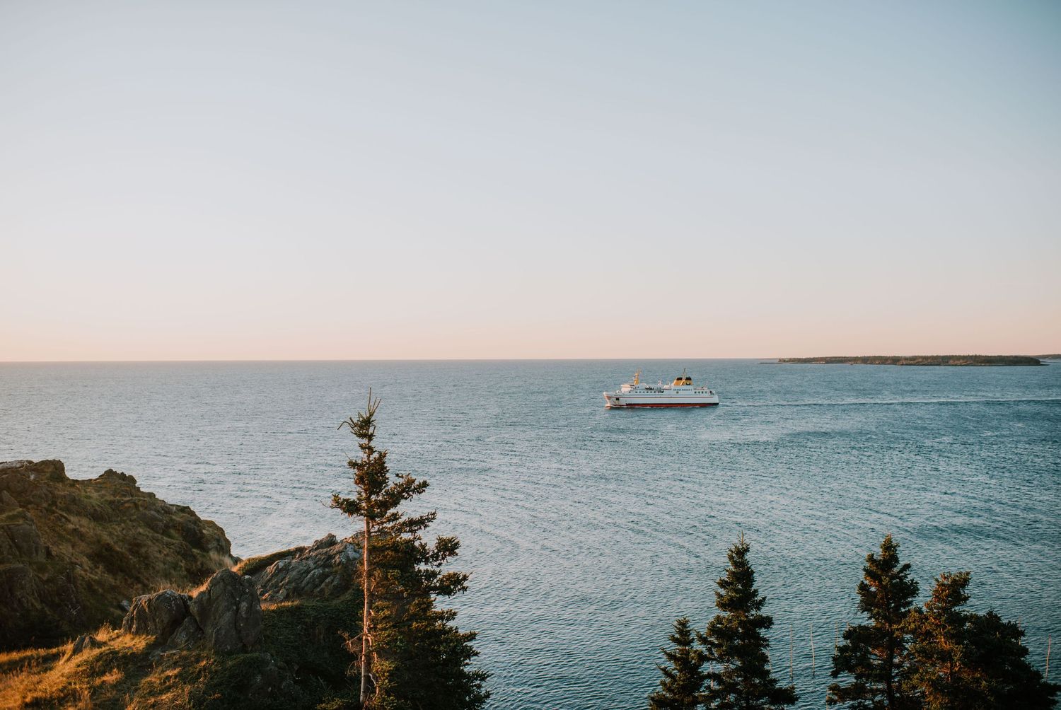 A white ferry boat sailing on calm blue ocean waters near rocky cliffs and evergreen trees at sunset.