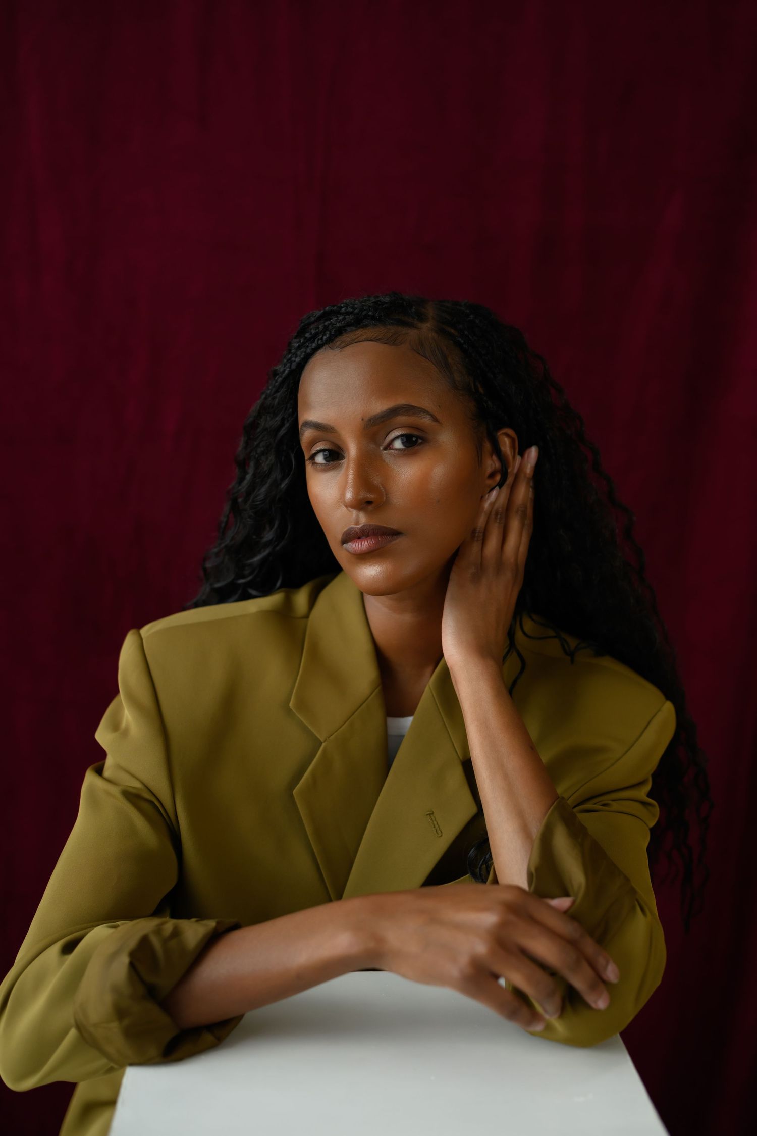 Young Black woman with long braids in olive blazer poses thoughtfully against deep red backdrop.