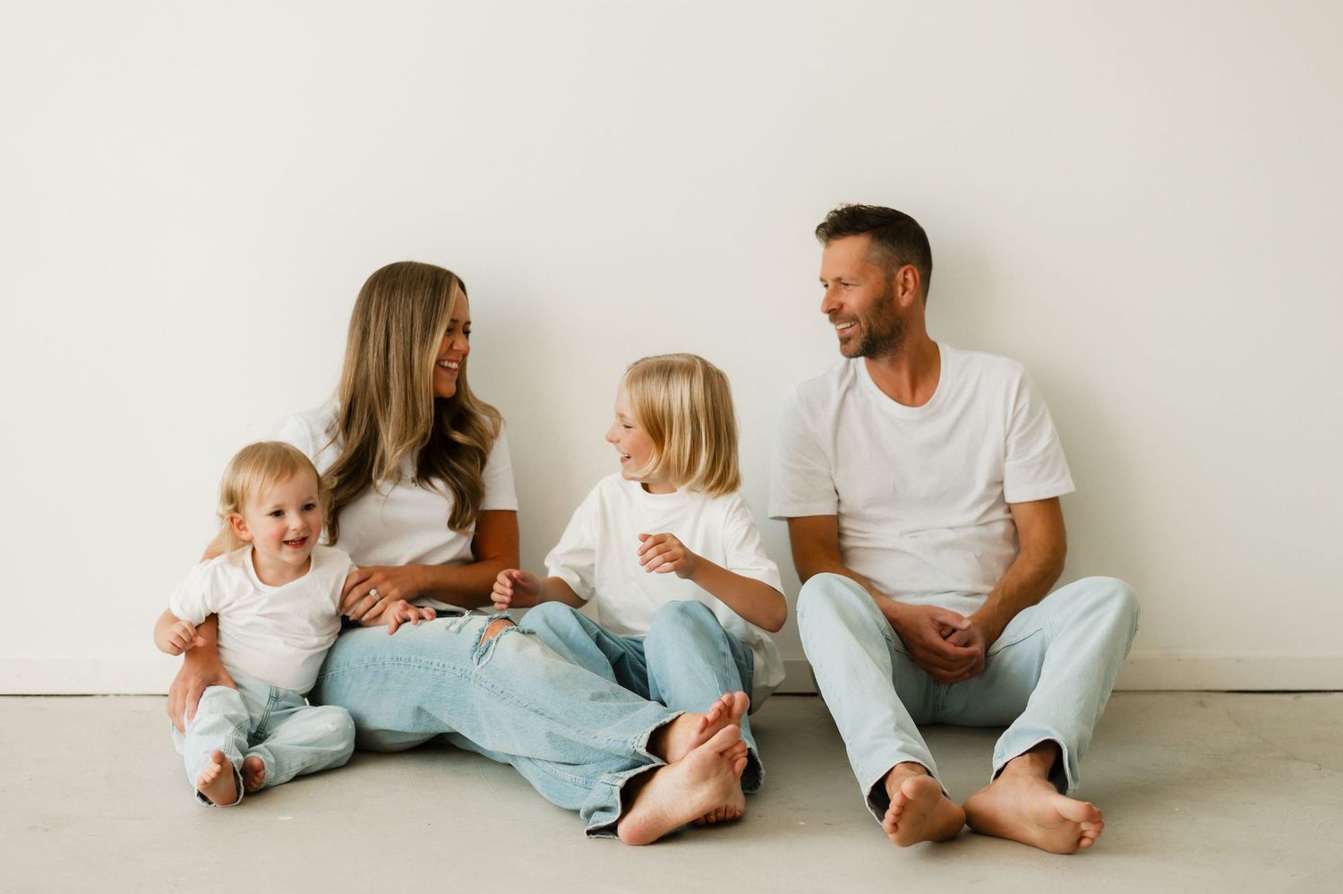 Happy family of four sitting on floor against white wall, parents smiling at children in casual white tops and jeans.