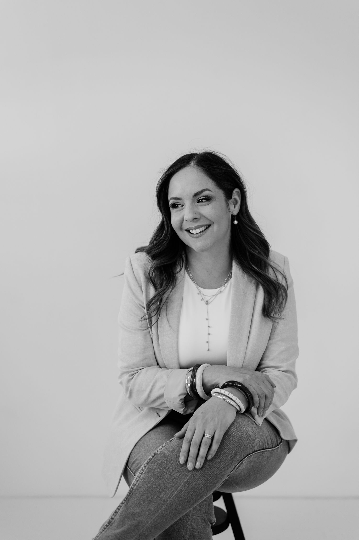 A woman in a white blazer sits casually and smiles while posing for a professional black and white portrait series.