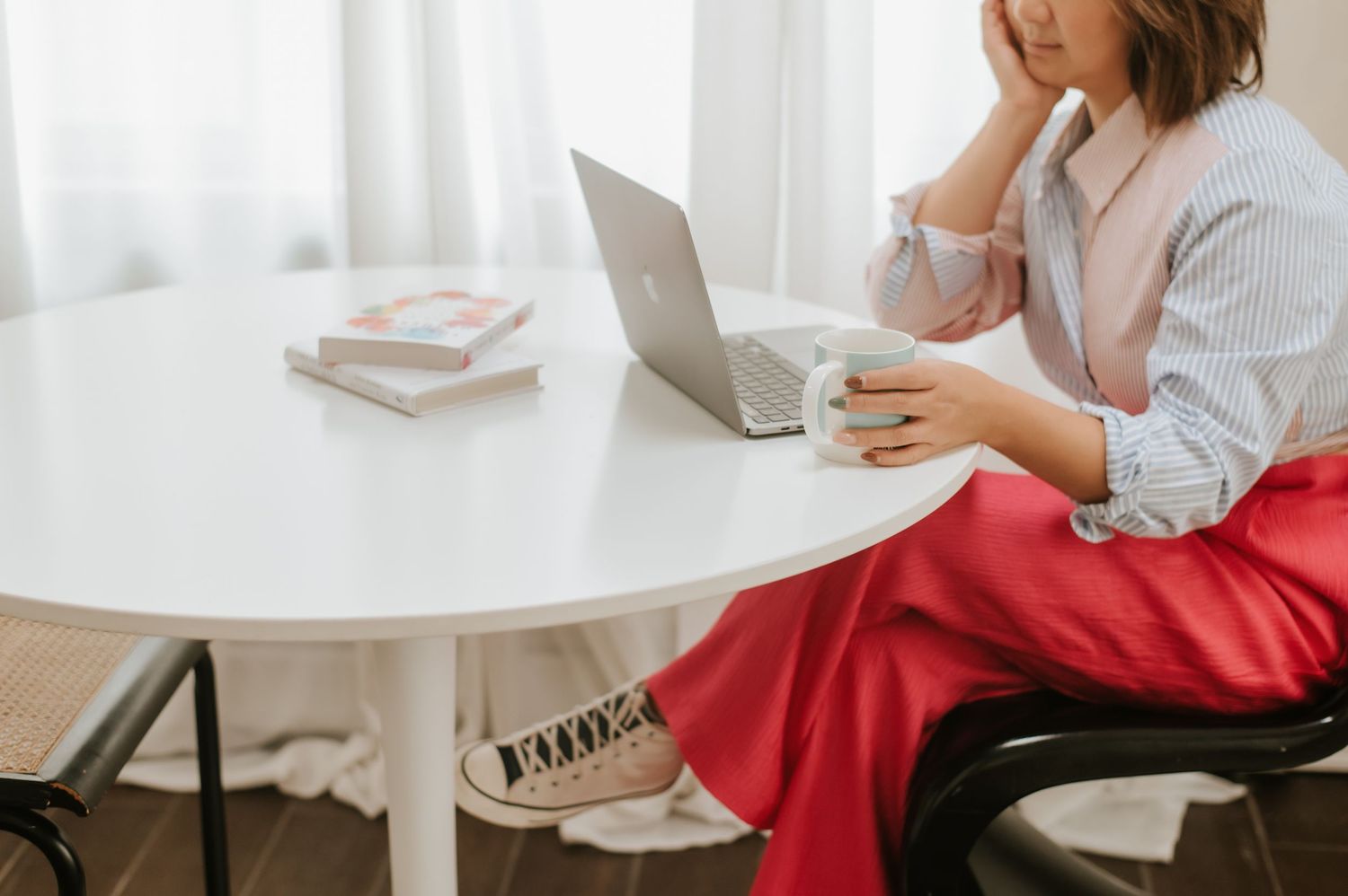 Person in a white sweater and red pants working on laptop while sitting at a round white table.