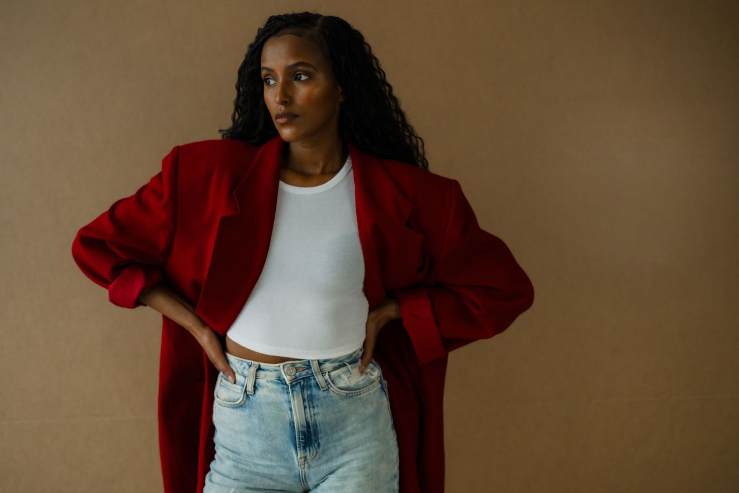 Woman in red blazer, white crop top, and light jeans posing confidently against a beige background.