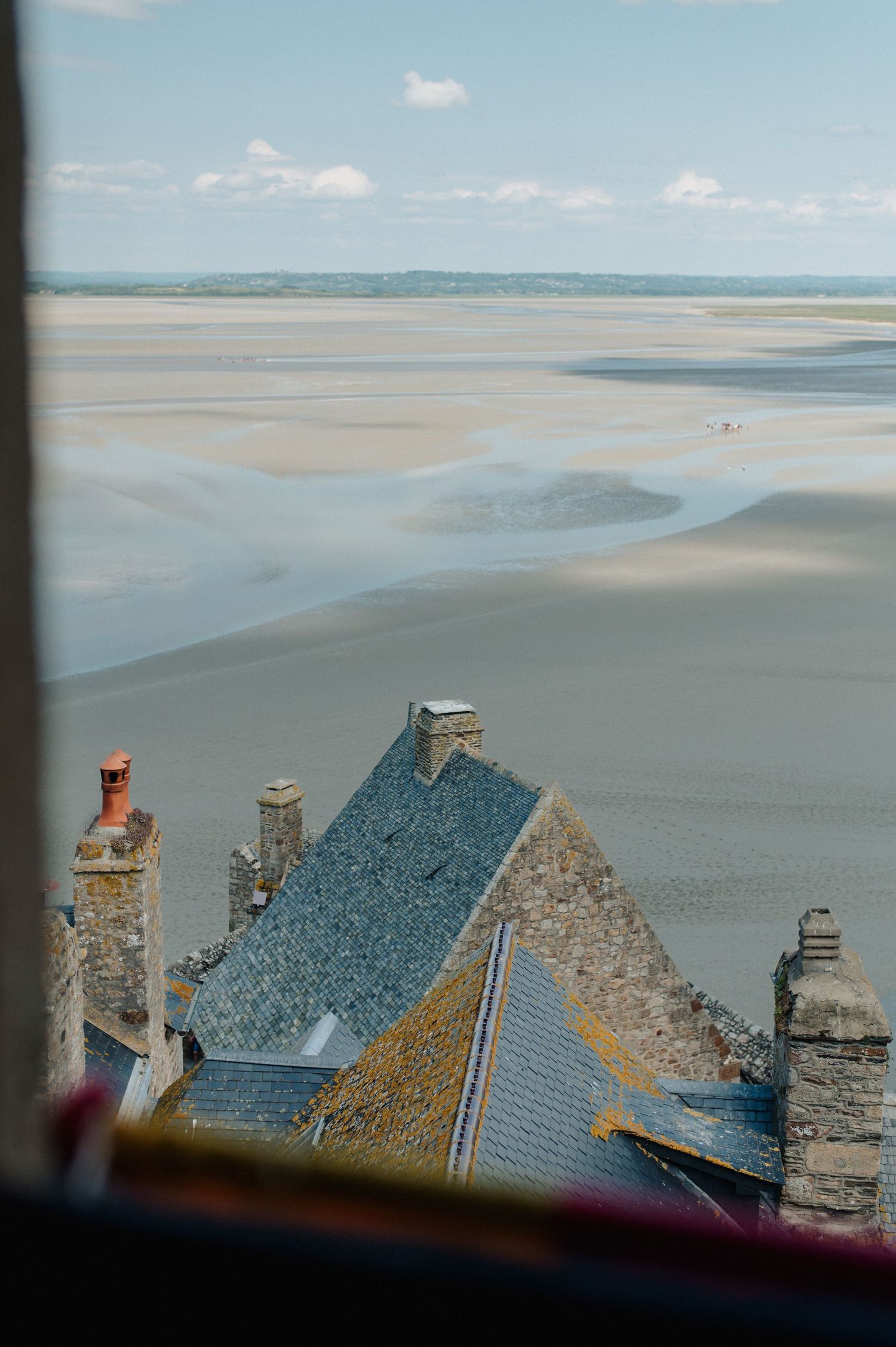 Aerial view of Mont Saint-Michel rooftops with vast tidal flats and bay stretching to the horizon under a cloudy sky.