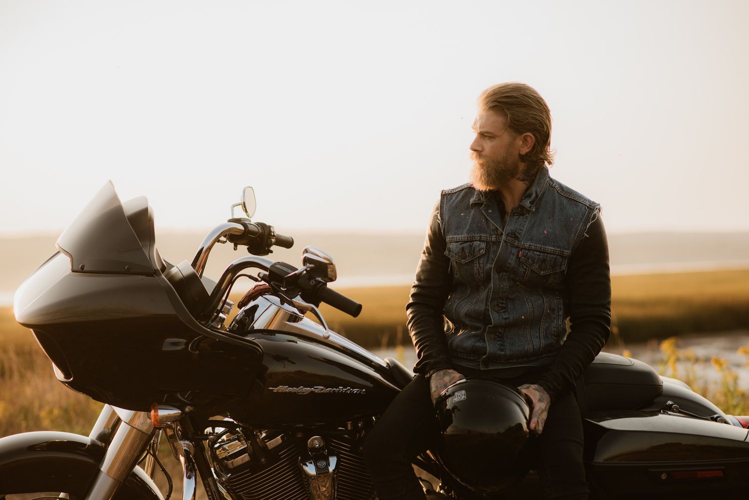 Man in denim jacket sitting on a Harley-Davidson motorcycle at sunset in an open field.