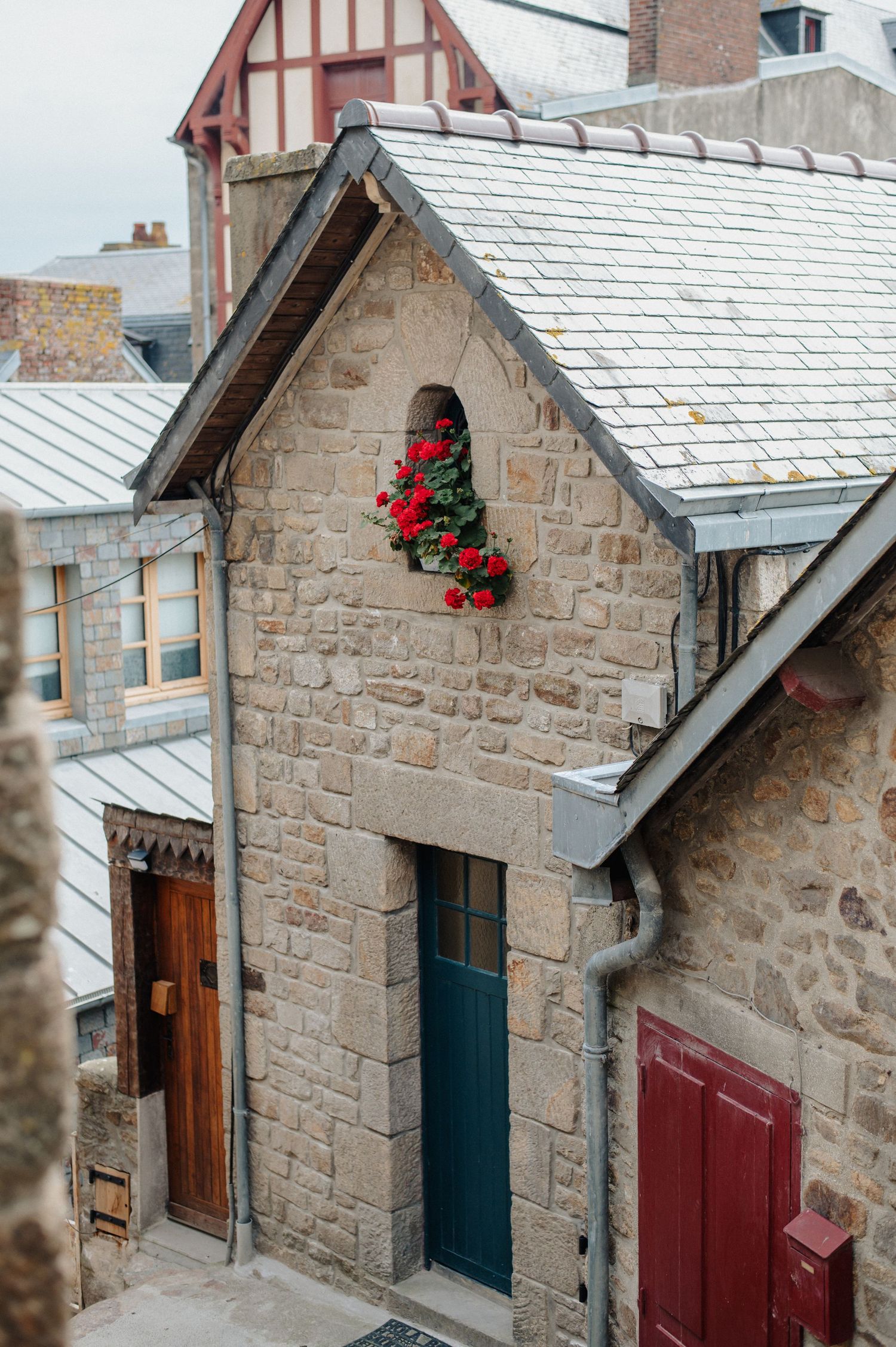 Stone building with red geraniums in window, slate roof, teal door in a charming French village.