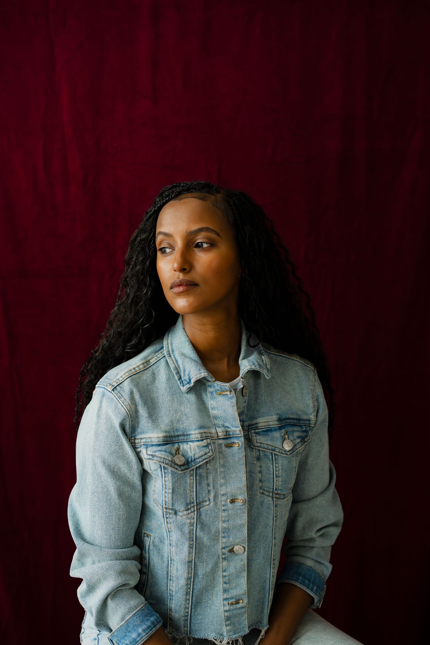 Young woman with long braids wearing a denim jacket poses against a deep red background.