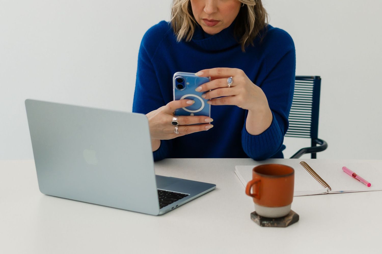 A person in a navy sweater works at a desk with a laptop, phone, and coffee mug.