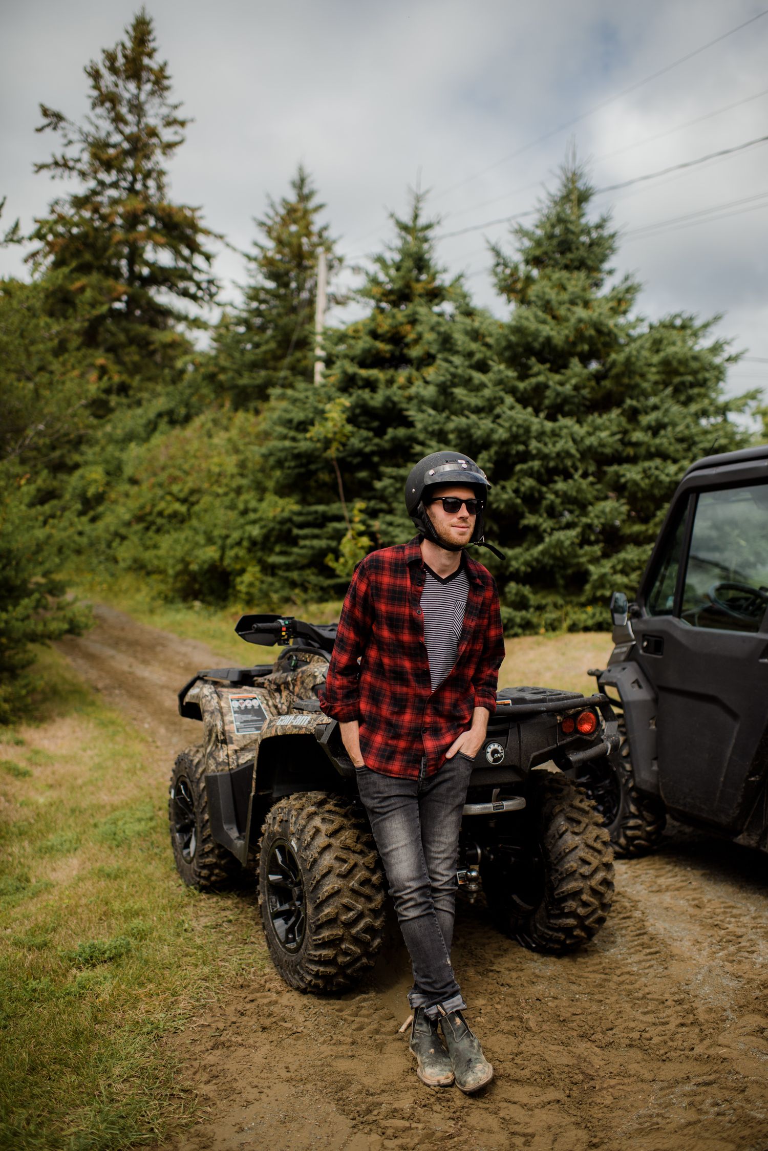 Man in plaid shirt and helmet leaning against a camouflage ATV in a forest trail.