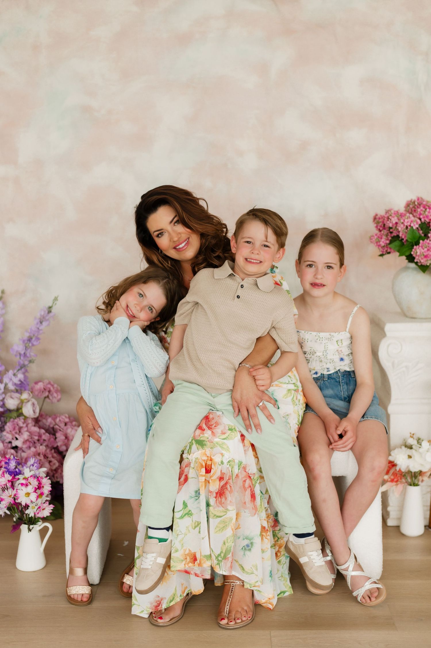 A smiling mother poses with her three young children in a bright studio with floral decor and pastel backdrop.