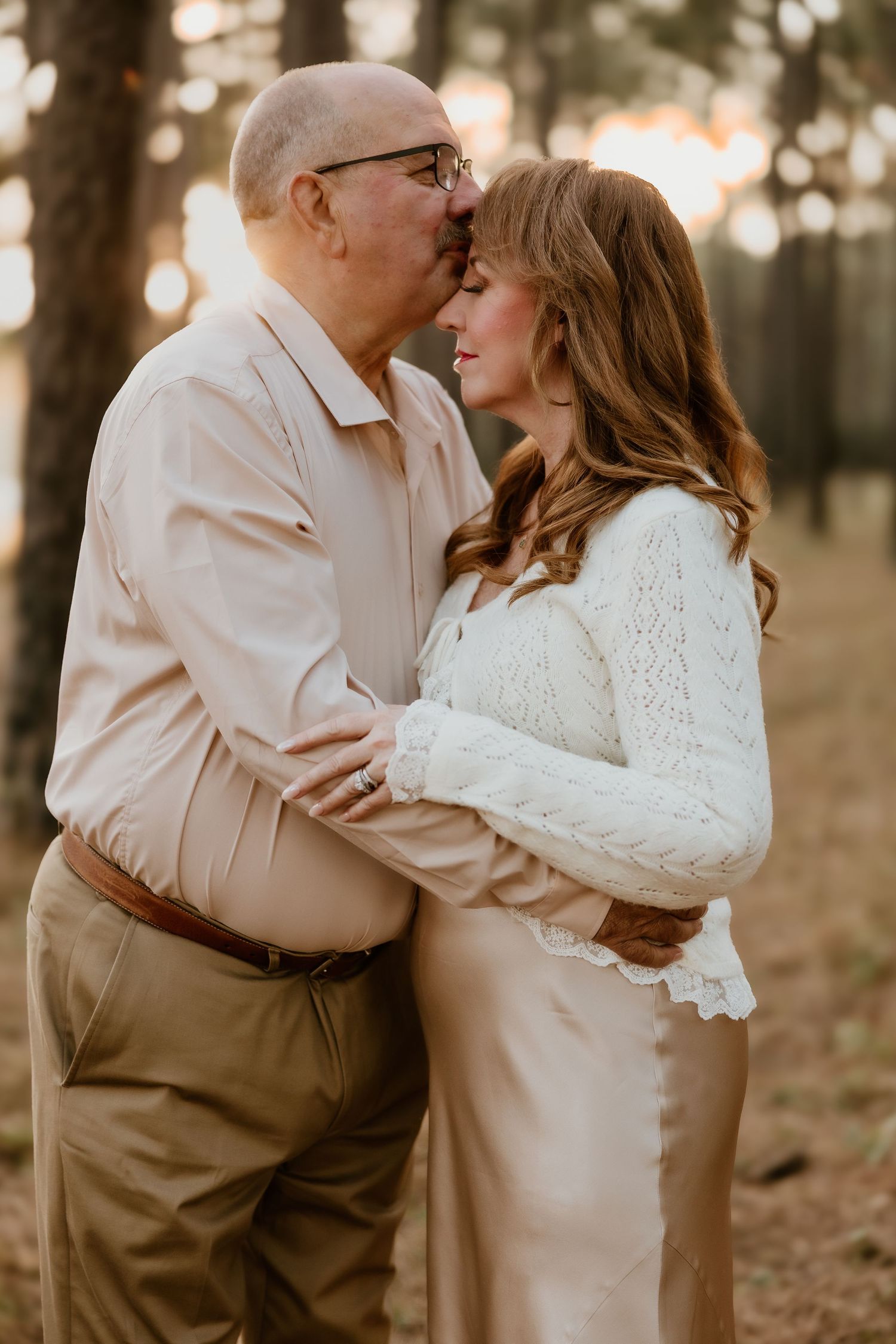 A romantic outdoor couple's portrait shows tender embrace and kiss in warm sunset lighting among trees.