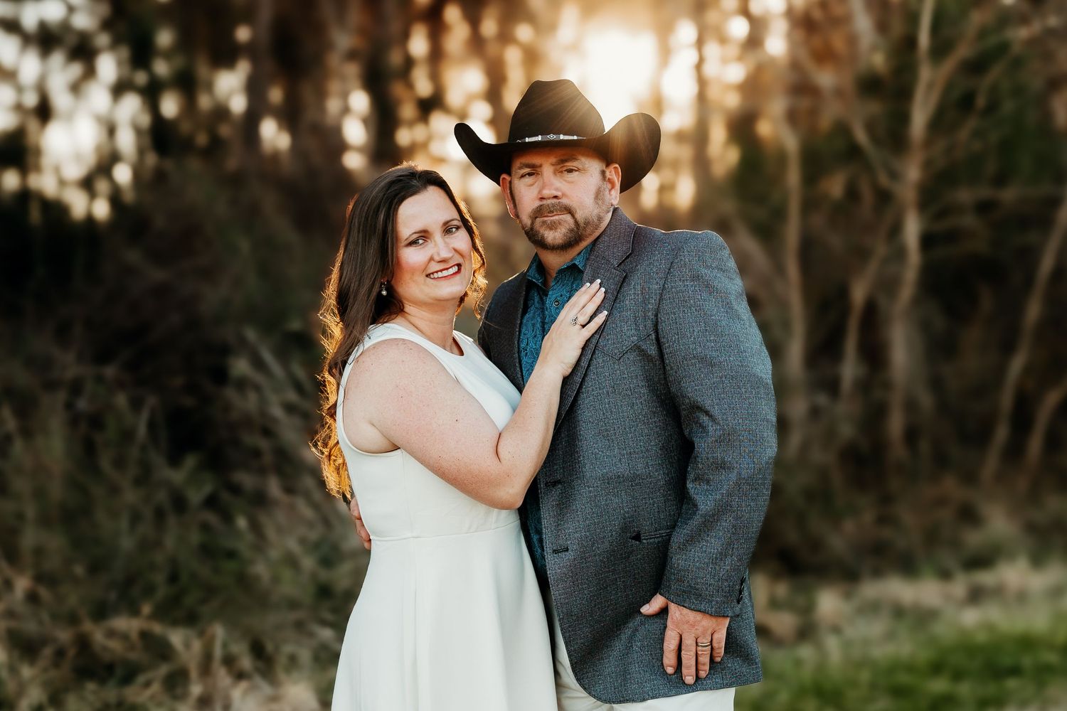 A couple poses together outdoors at sunset, with the person in a white dress and their partner wearing a cowboy hat and blazer.