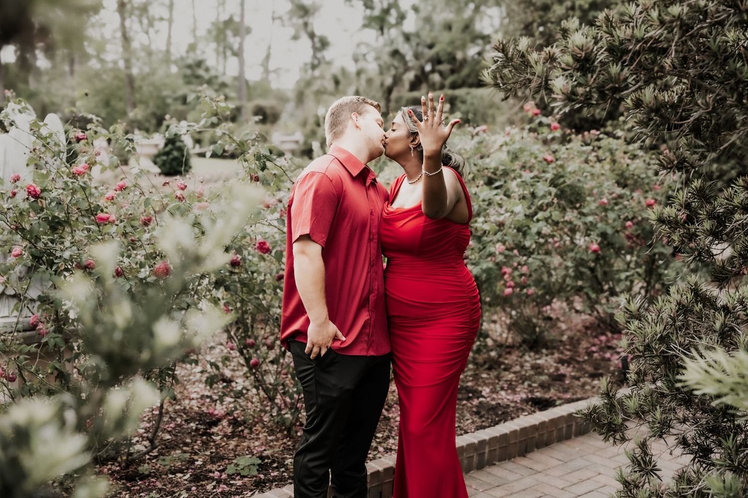 Couple in matching red outfits sharing a kiss surrounded by lush greenery and red roses in a garden setting.
