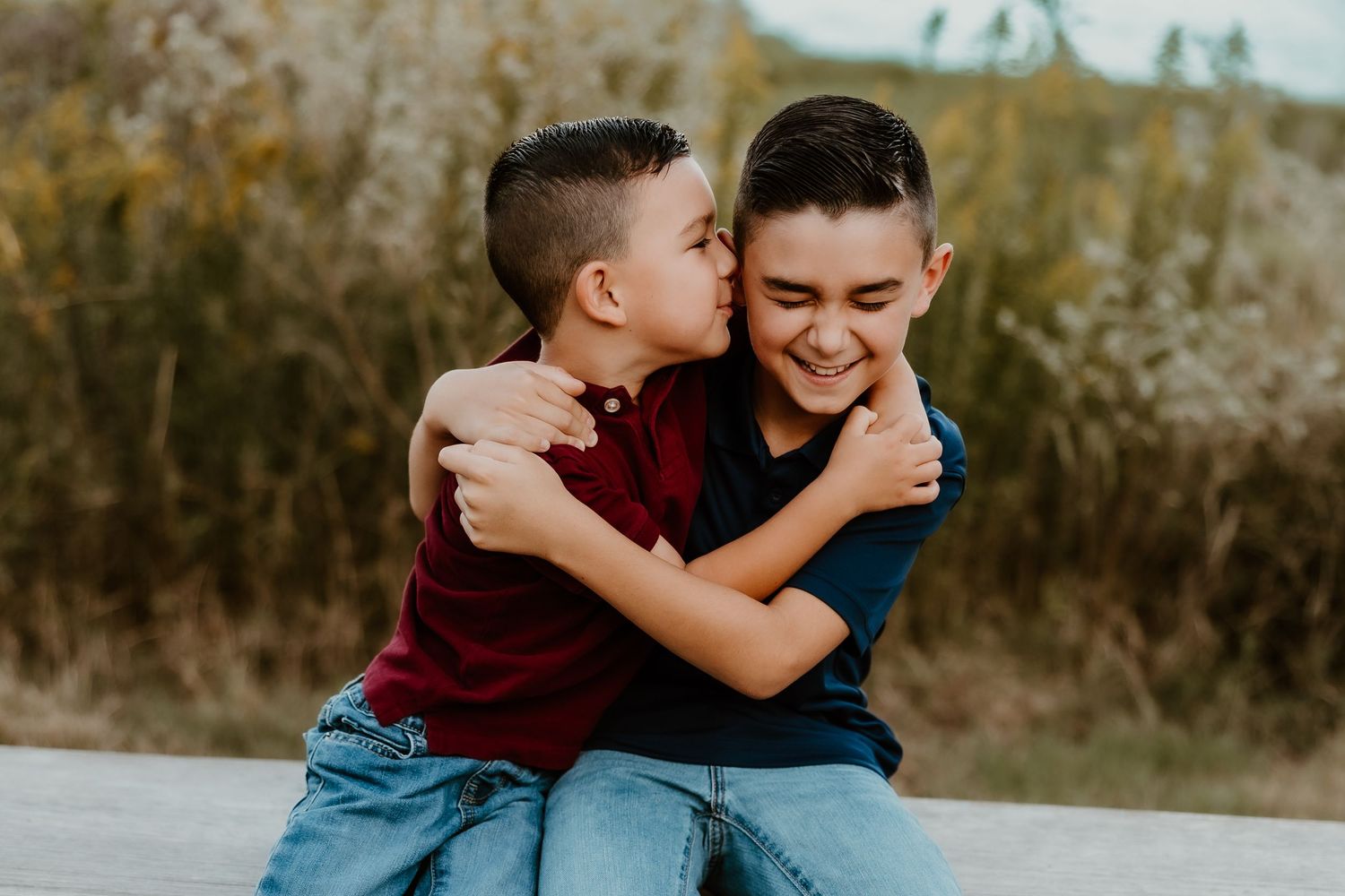 Two brothers laughing and embracing while sitting on a rustic wooden fence.