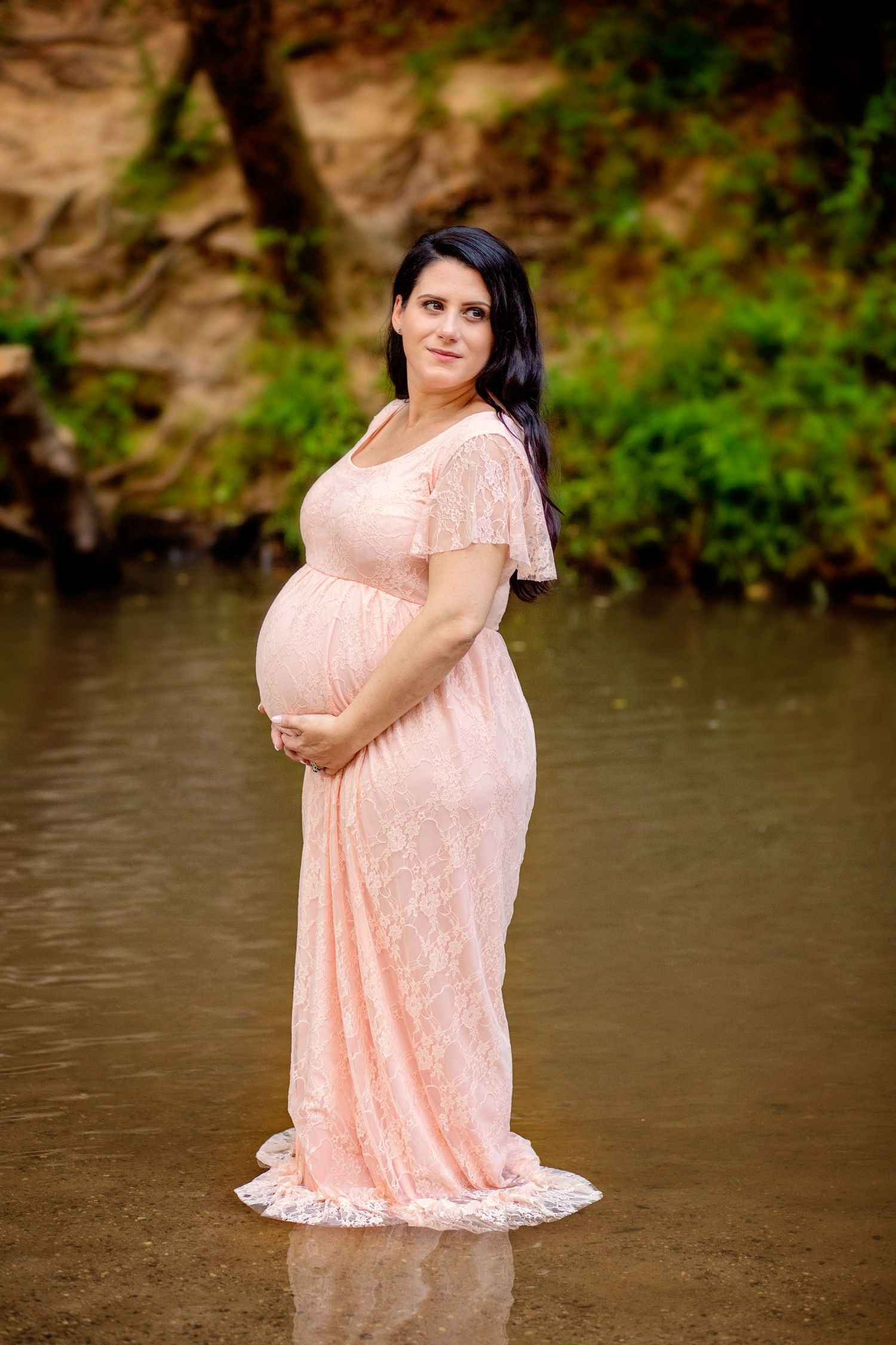 A woman in a flowing peach lace dress poses by a waterfront during a photo session.