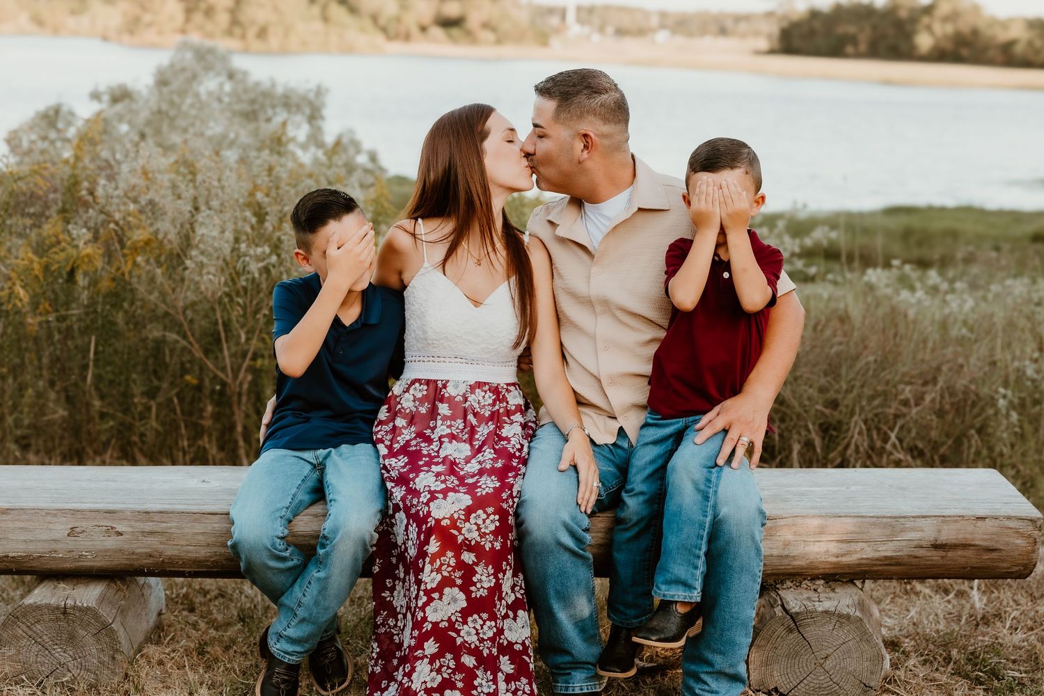 A family sits playfully on a wooden bench near a lake during sunset.