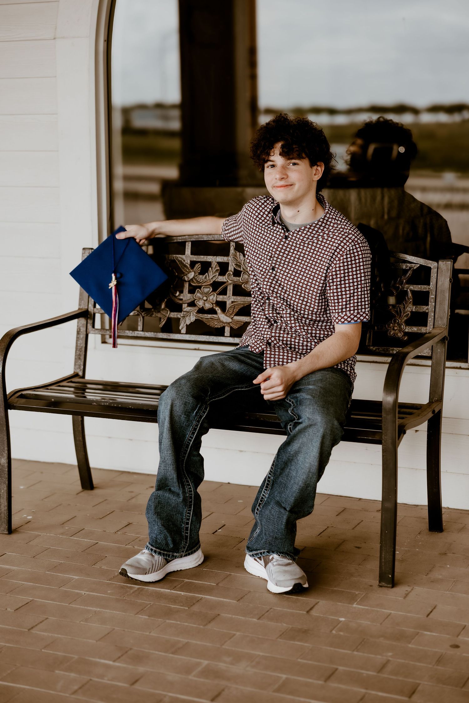 Young man in checkered shirt holding graduation cap while sitting on decorative bench outdoors.