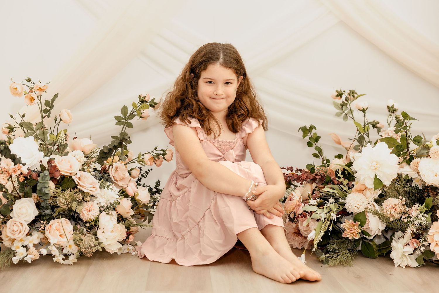 Young girl in pink dress sitting among pastel floral arrangements in a bright studio portrait setting.
