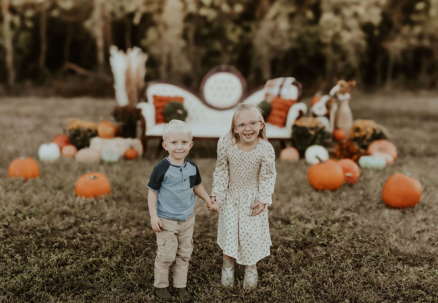 Two children stand together in a fall garden setting with pumpkins and white furniture in the background.