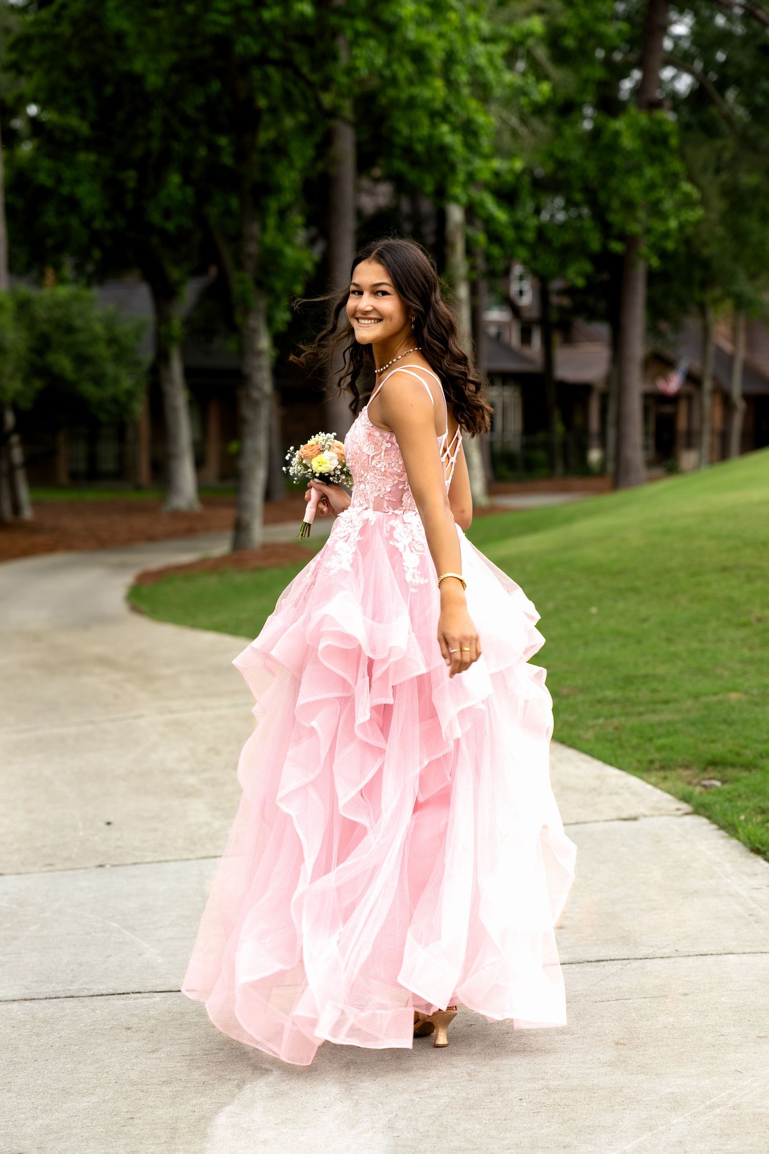 Young woman in pink ruffled prom gown holding bouquet, posing on pathway surrounded by trees.