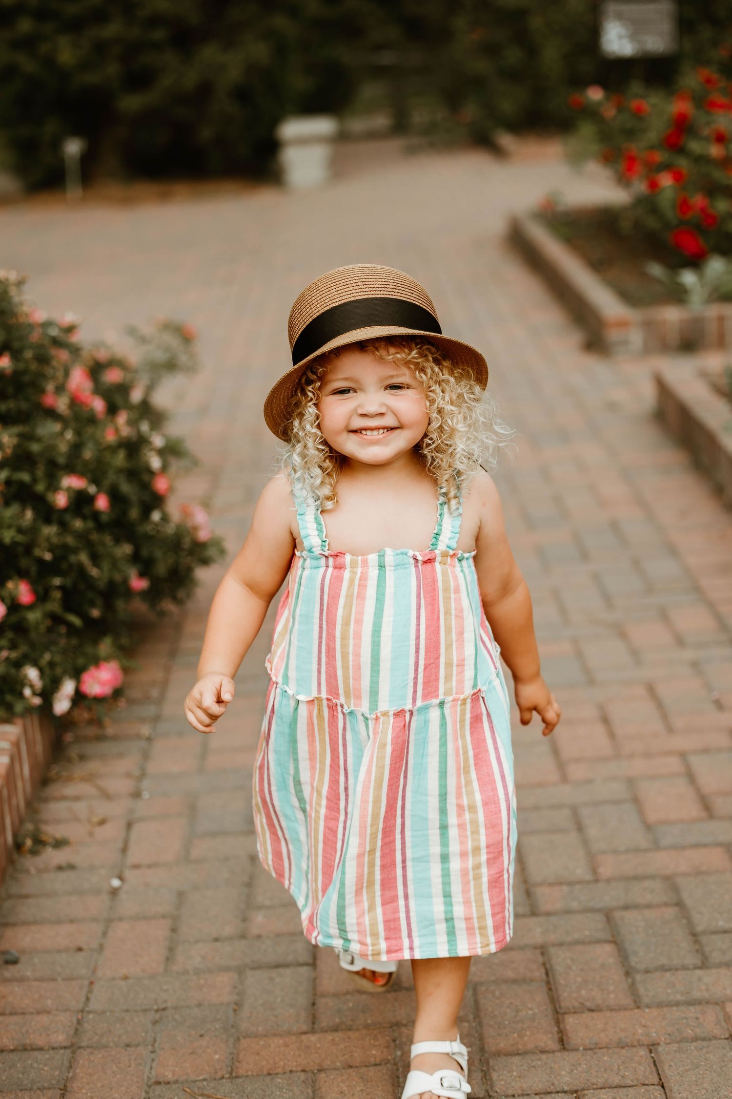 A young child in a striped summer dress and straw hat plays on a brick patio surrounded by blooming rose bushes.