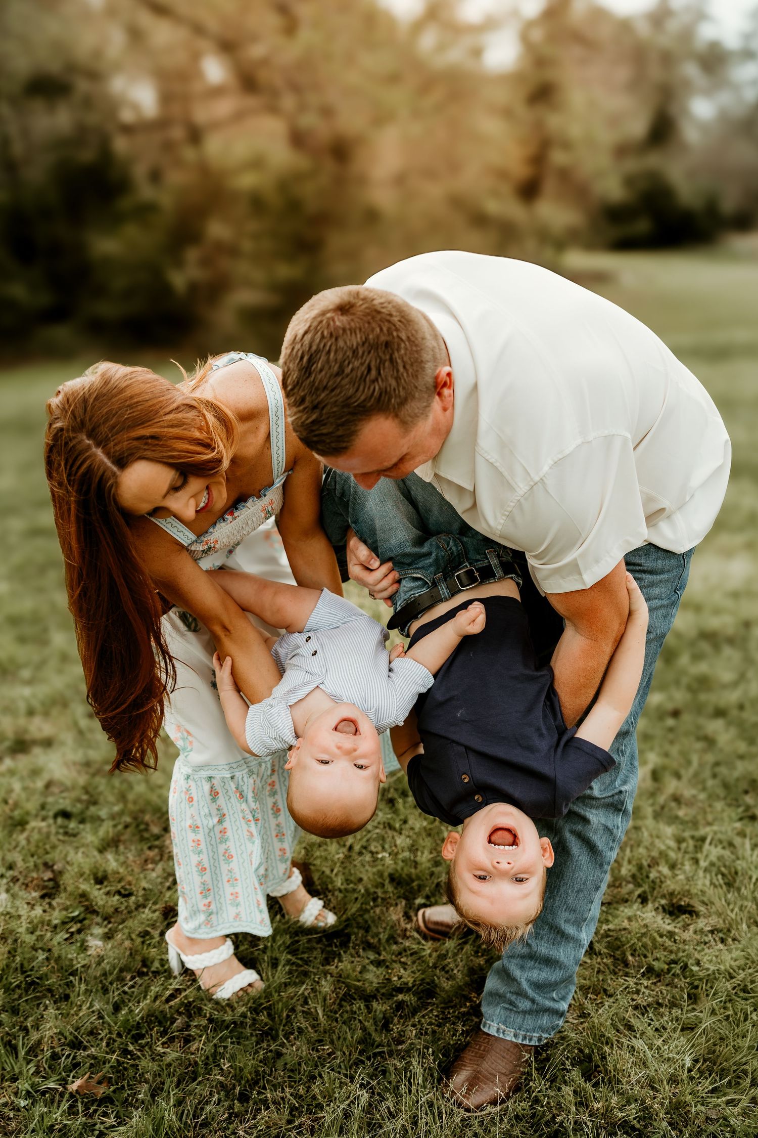 Playful family moment outdoors as parents hold two laughing children upside down on grassy lawn at sunset.