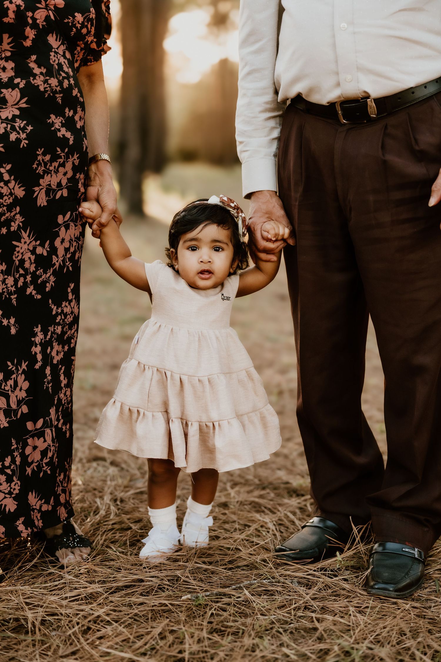 A toddler in a white dress stands between adults wearing brown and floral clothing during an outdoor photoshoot.