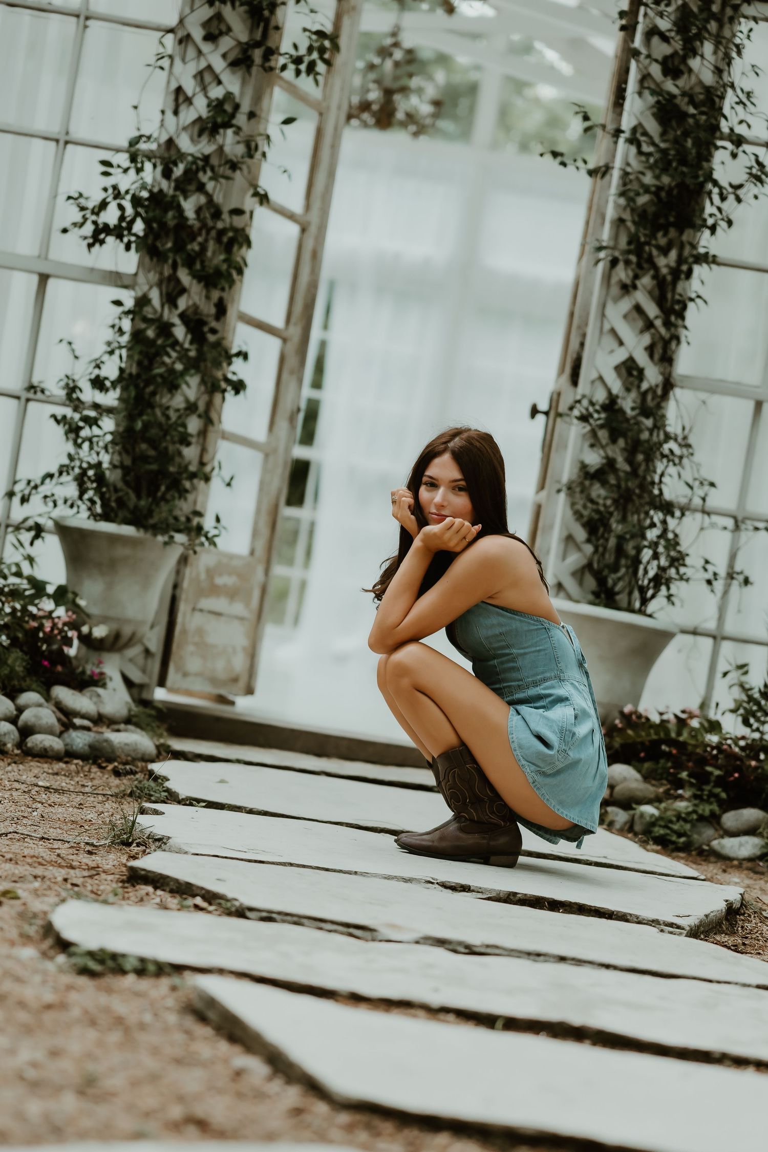 A model in a blue dress crouches on wooden planks surrounded by climbing vines.