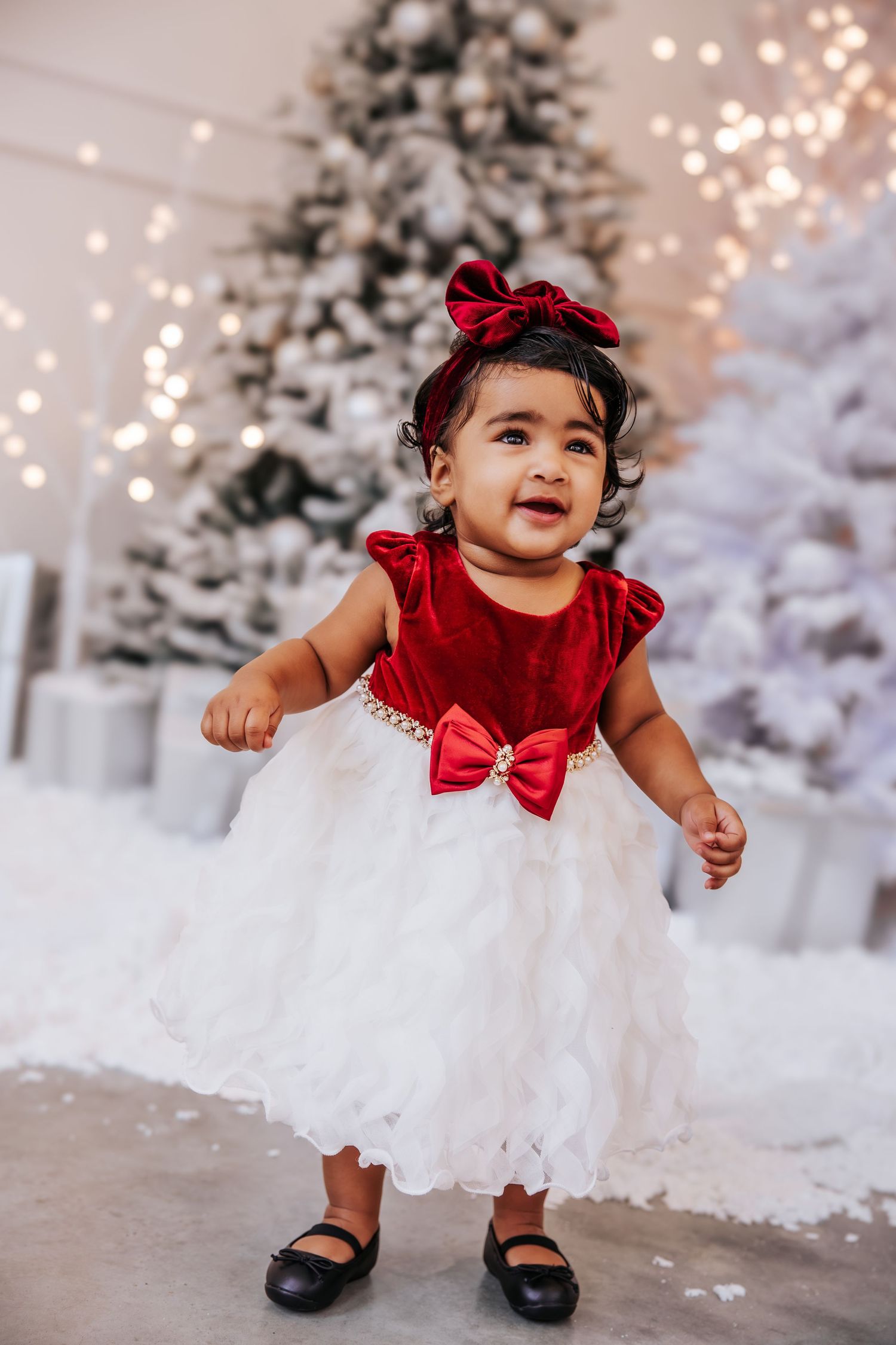 A baby in a white tulle dress with red bow sits in a winter wonderland setting with snowy trees and twinkling lights.
