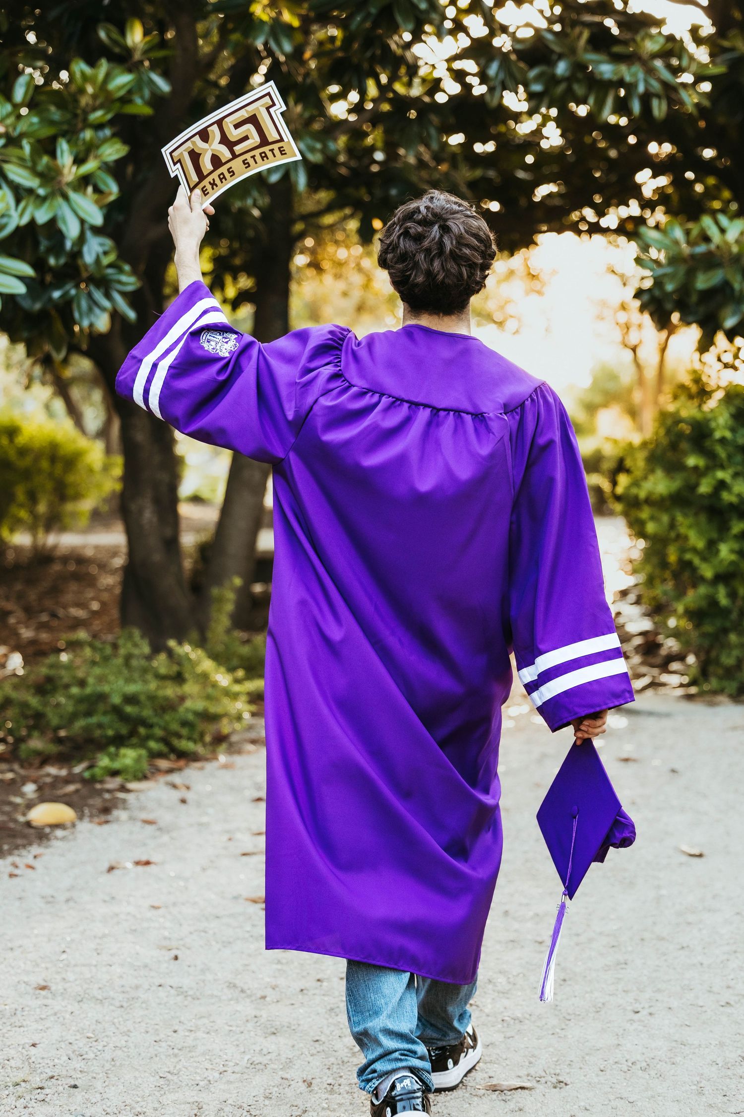 Person in purple graduation gown and jeans walking away on outdoor path.