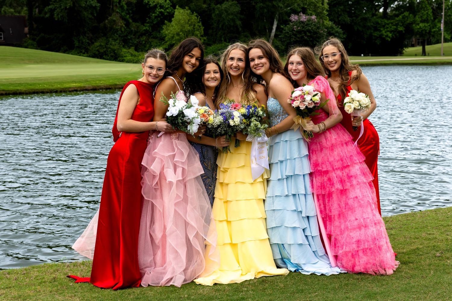 Seven bridesmaids in colorful rainbow gowns holding bouquets pose together near a pond at an outdoor wedding.
