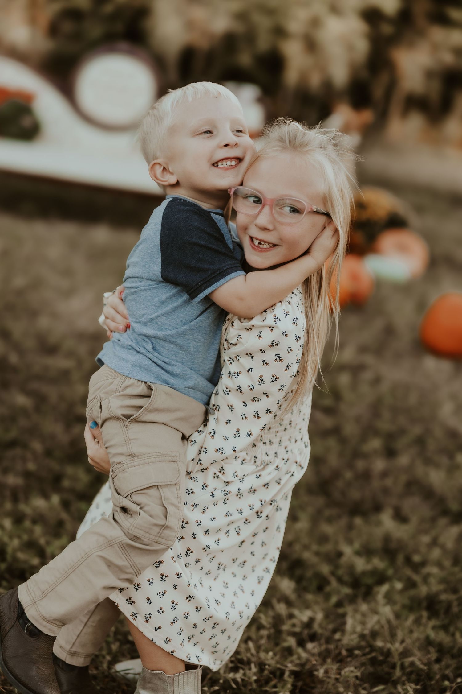 Two children share a joyful embrace while standing in a field with pumpkins scattered around.