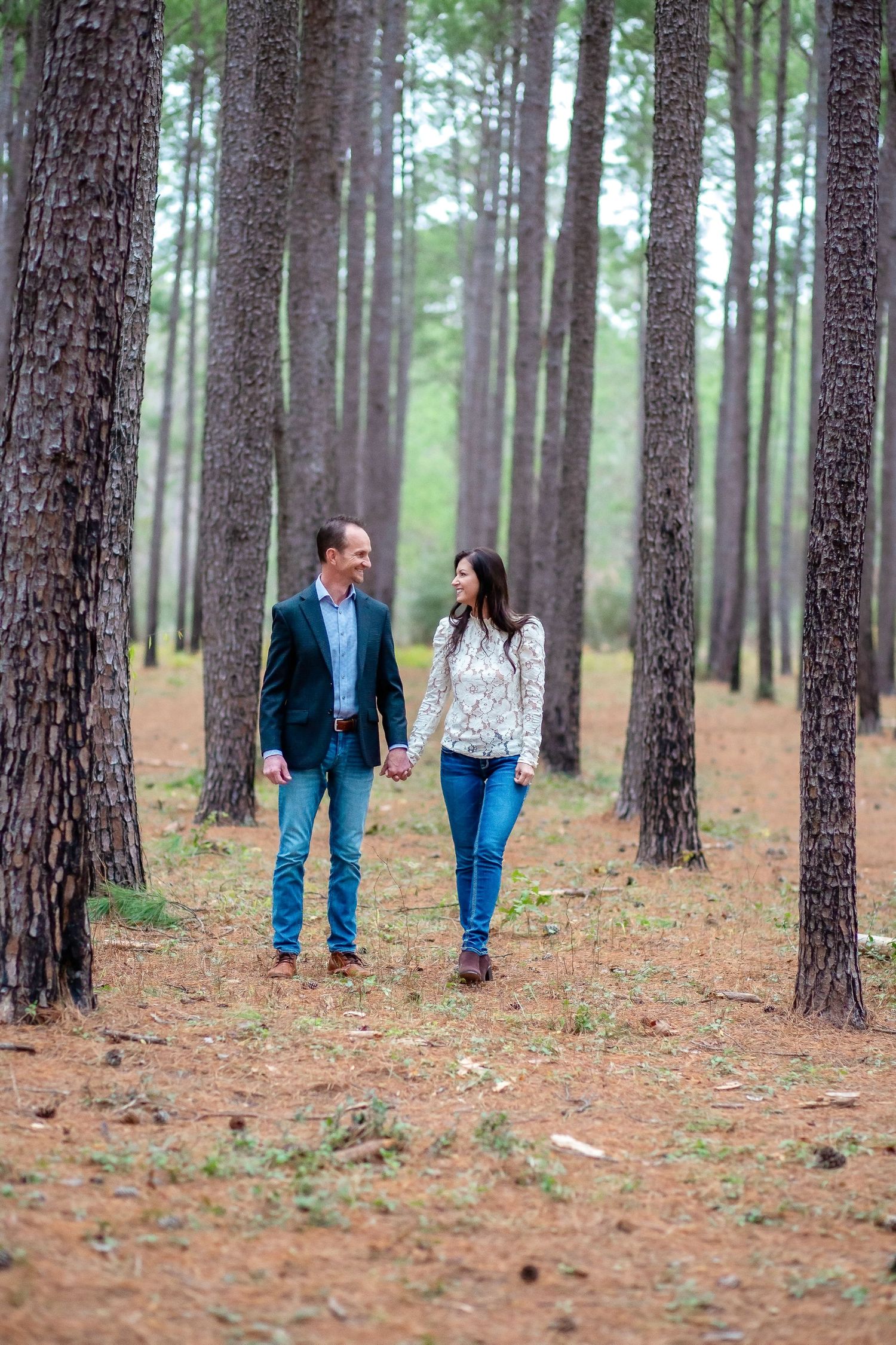 Couple walking hand in hand through pine forest during engagement session.