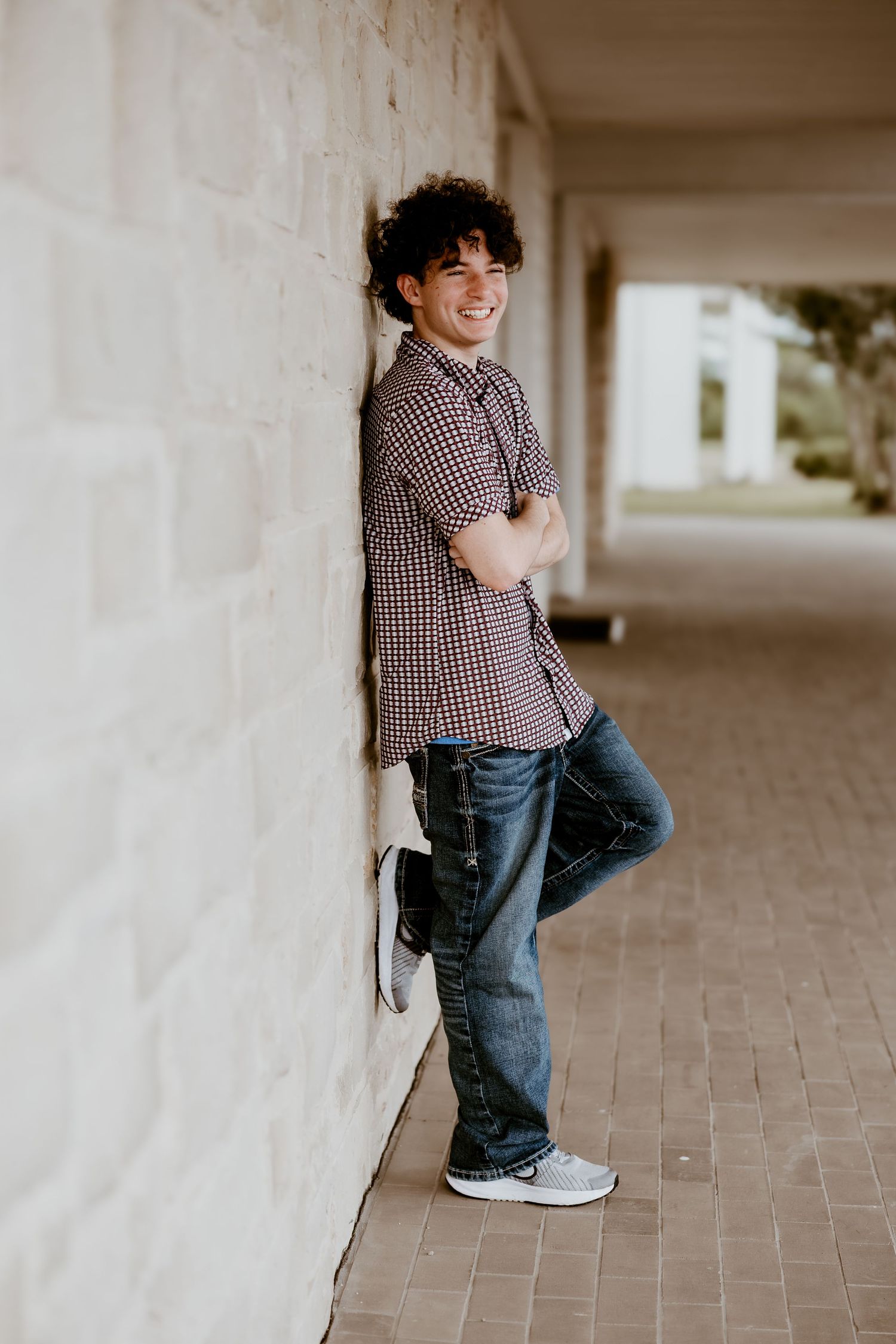 Smiling young man with curly hair leaning against a brick wall in a plaid shirt and jeans.
