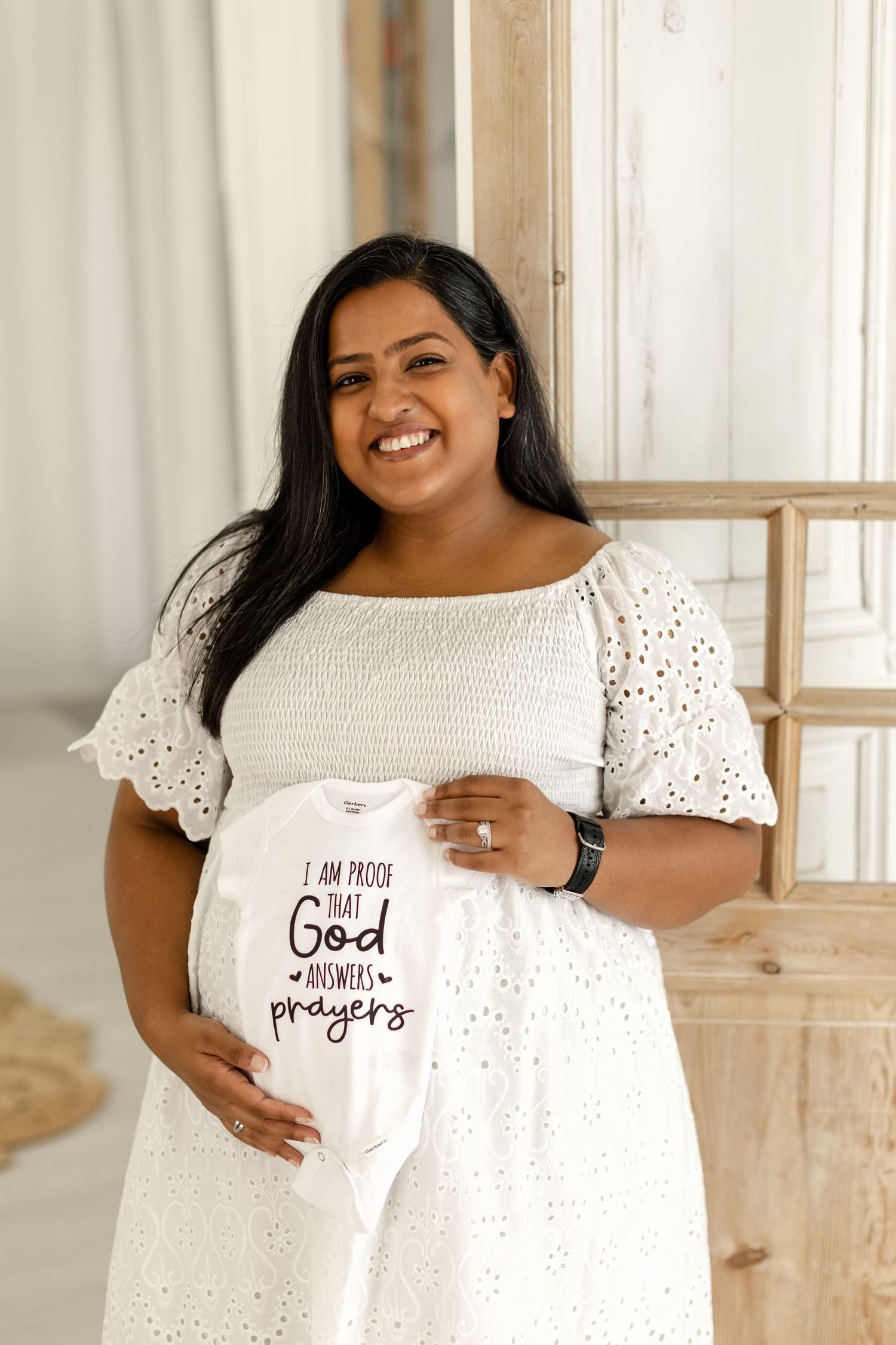 Smiling pregnant woman in white eyelet dress holding a baby onesie that reads I Am Proof That God Answers Prayers.