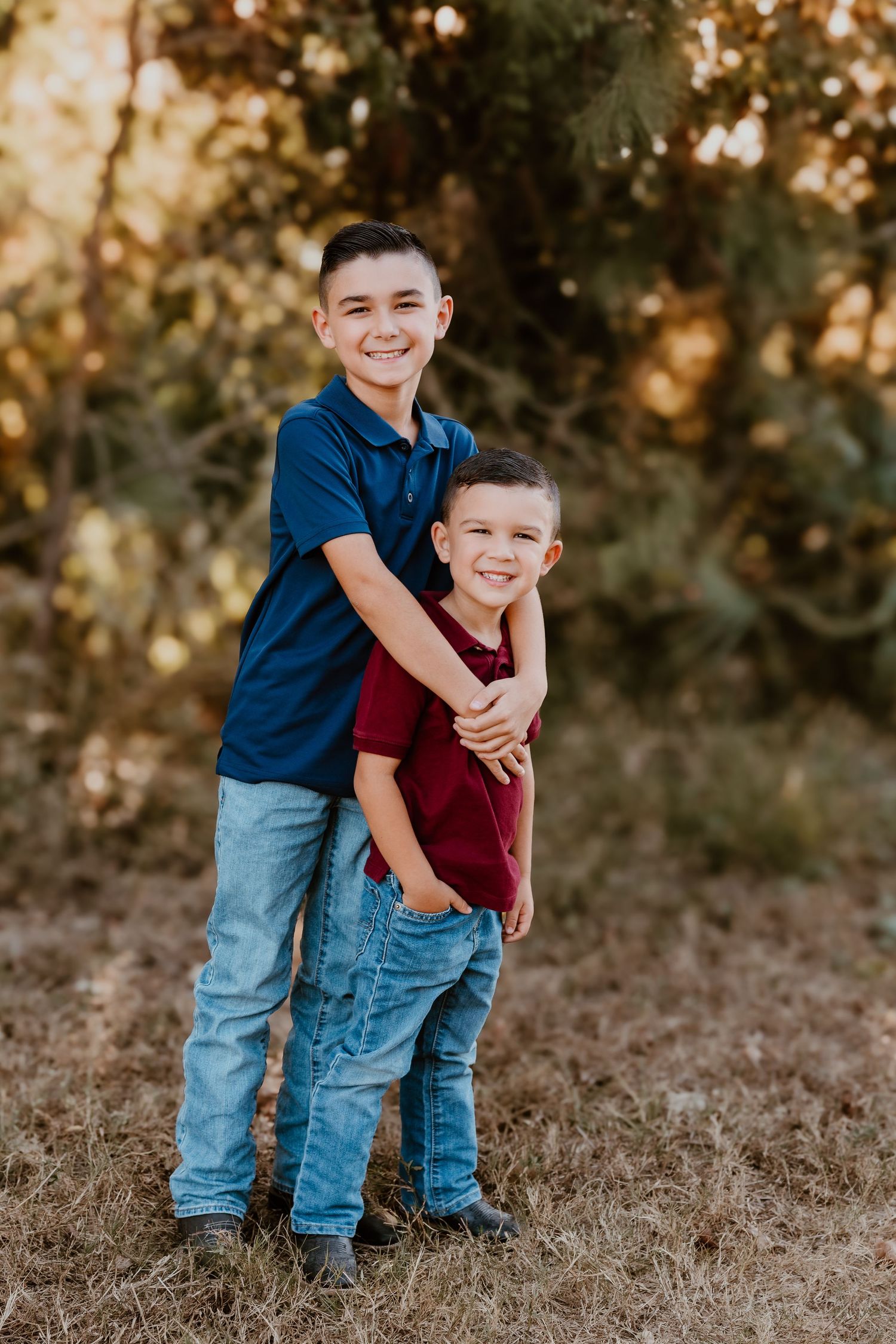 Two boys in blue and burgundy shirts embrace while posing outdoors.
