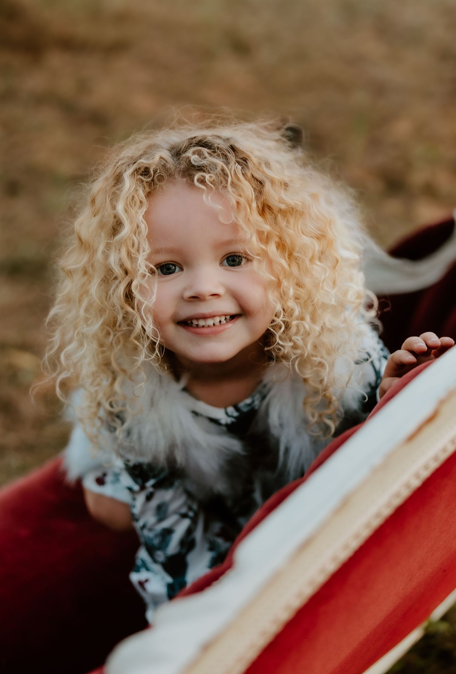 A small child with wild blonde curls smiles while sitting in a red wagon outdoors.