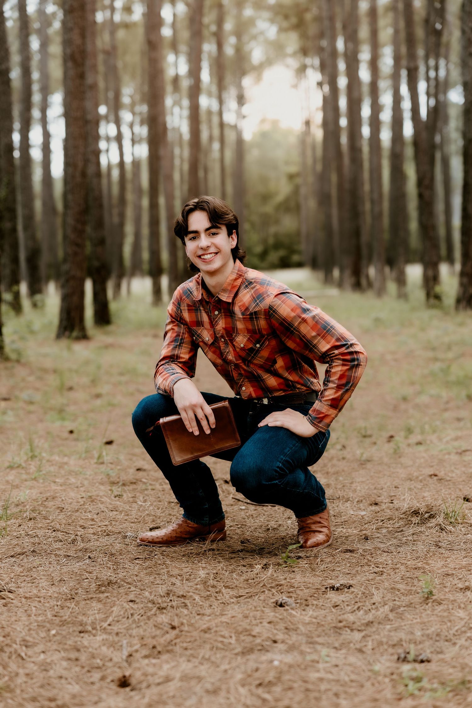 Young man in plaid shirt crouching in pine forest, smiling and holding a brown leather book outdoors.