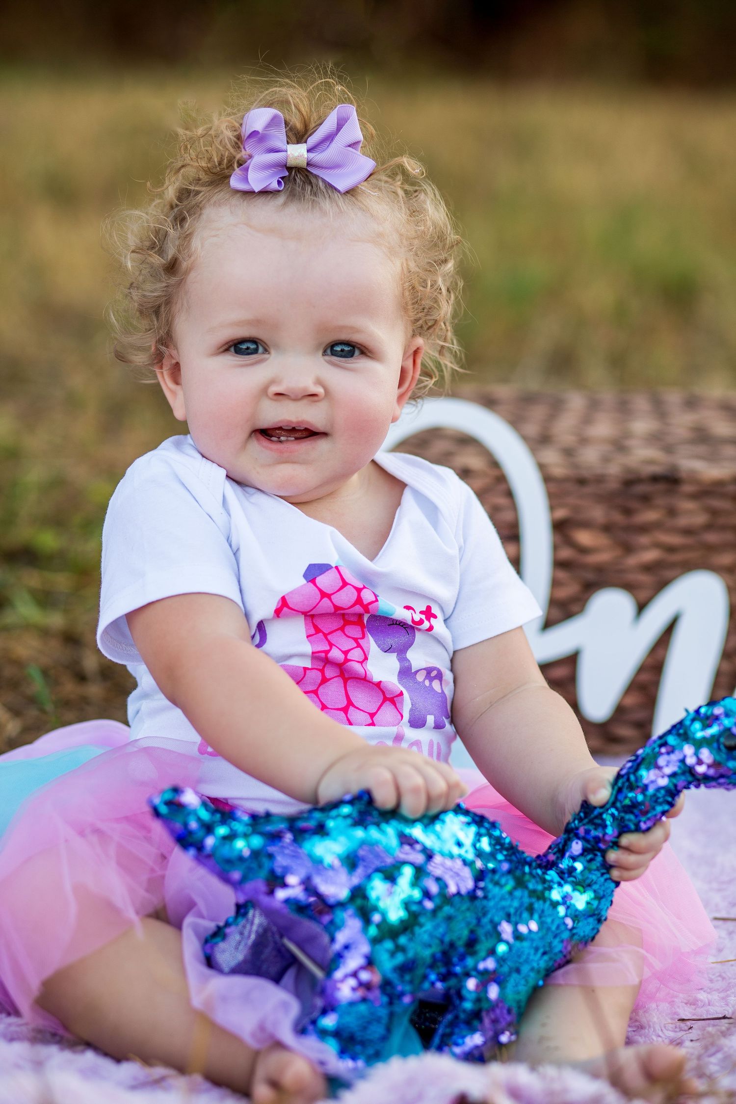 Baby wearing white dress and purple bow sits in sparkly blue and pink tutu dress.