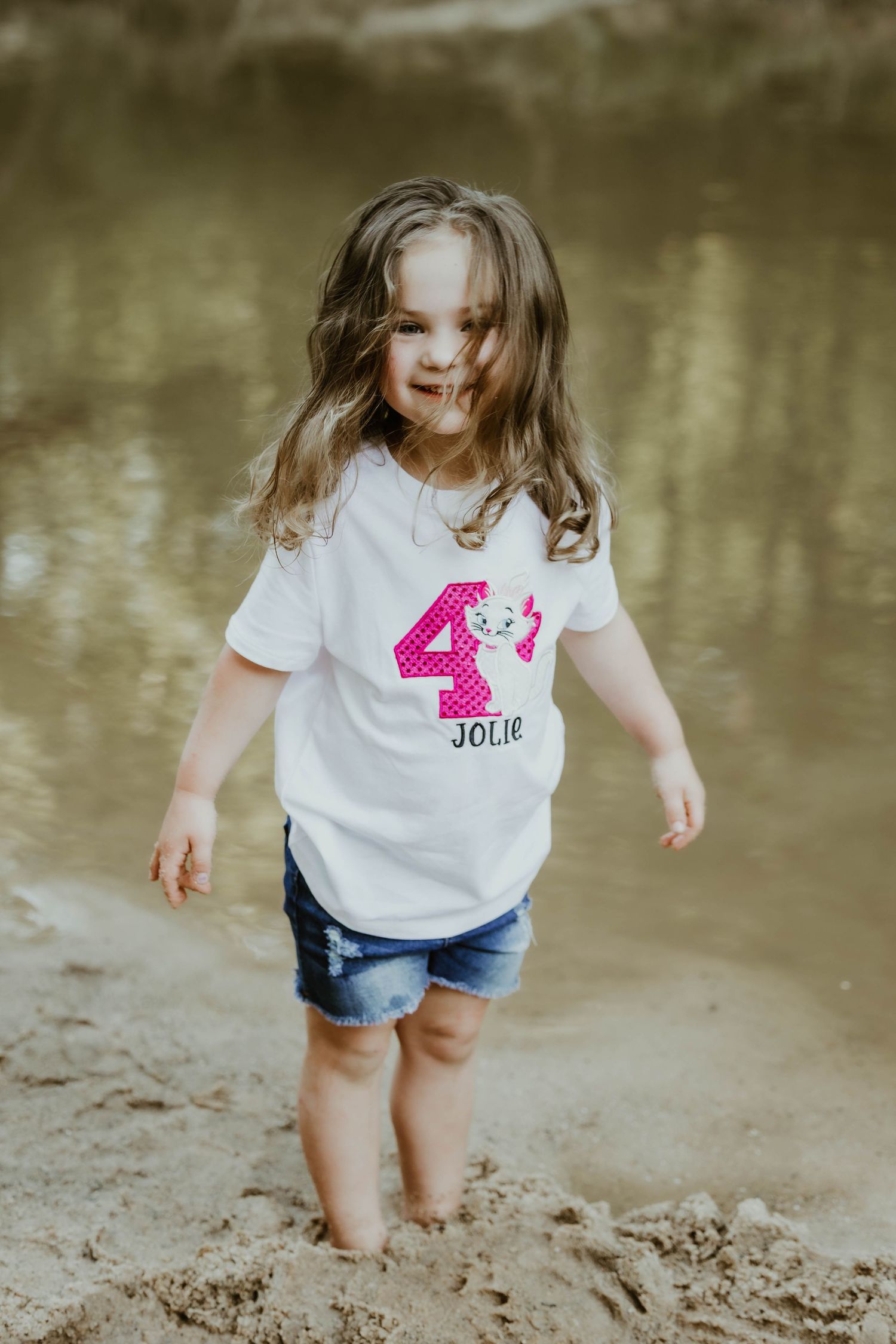 Child wearing birthday shirt plays happily along creek shoreline.