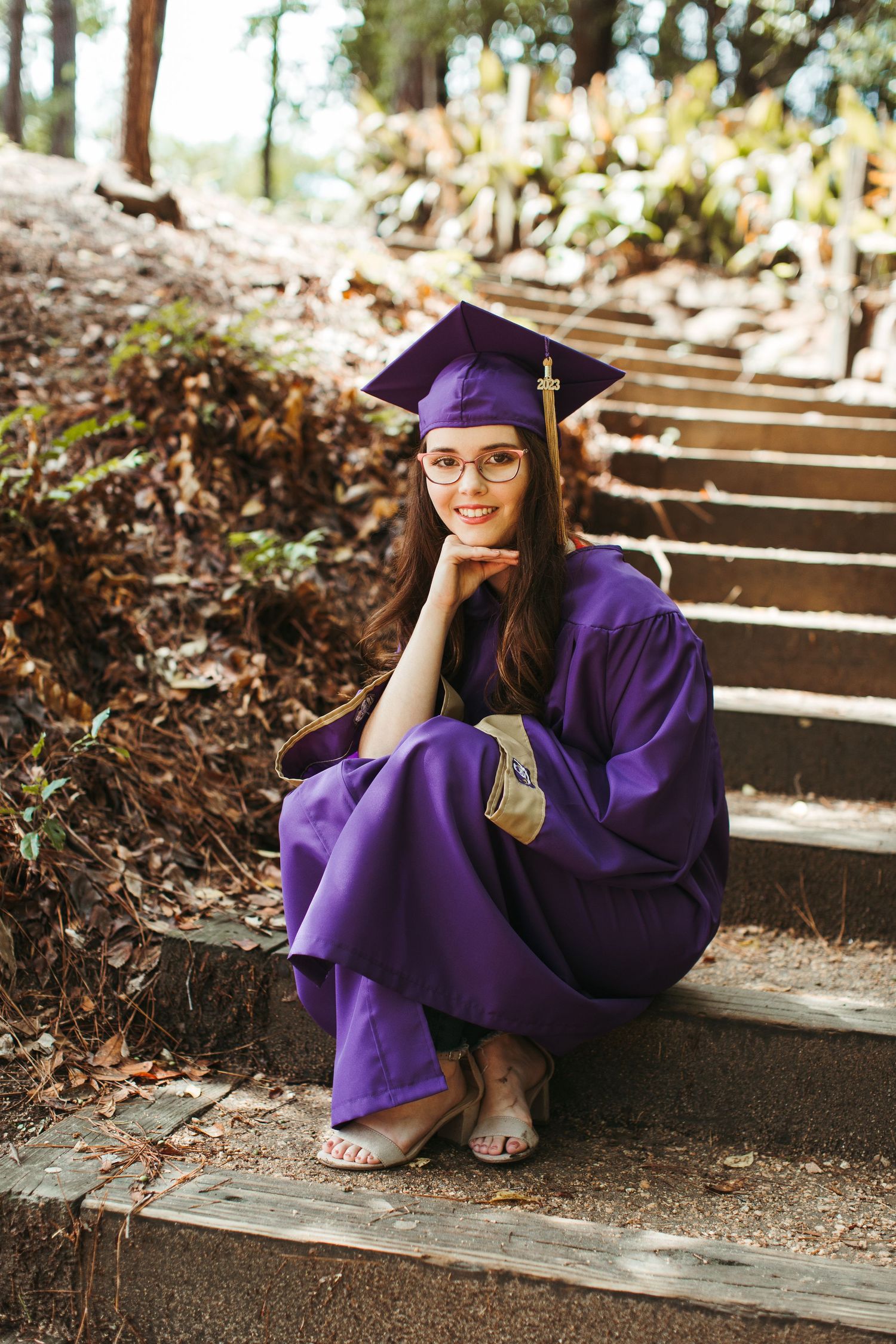Graduate in purple regalia sitting on outdoor stone steps surrounded by foliage.