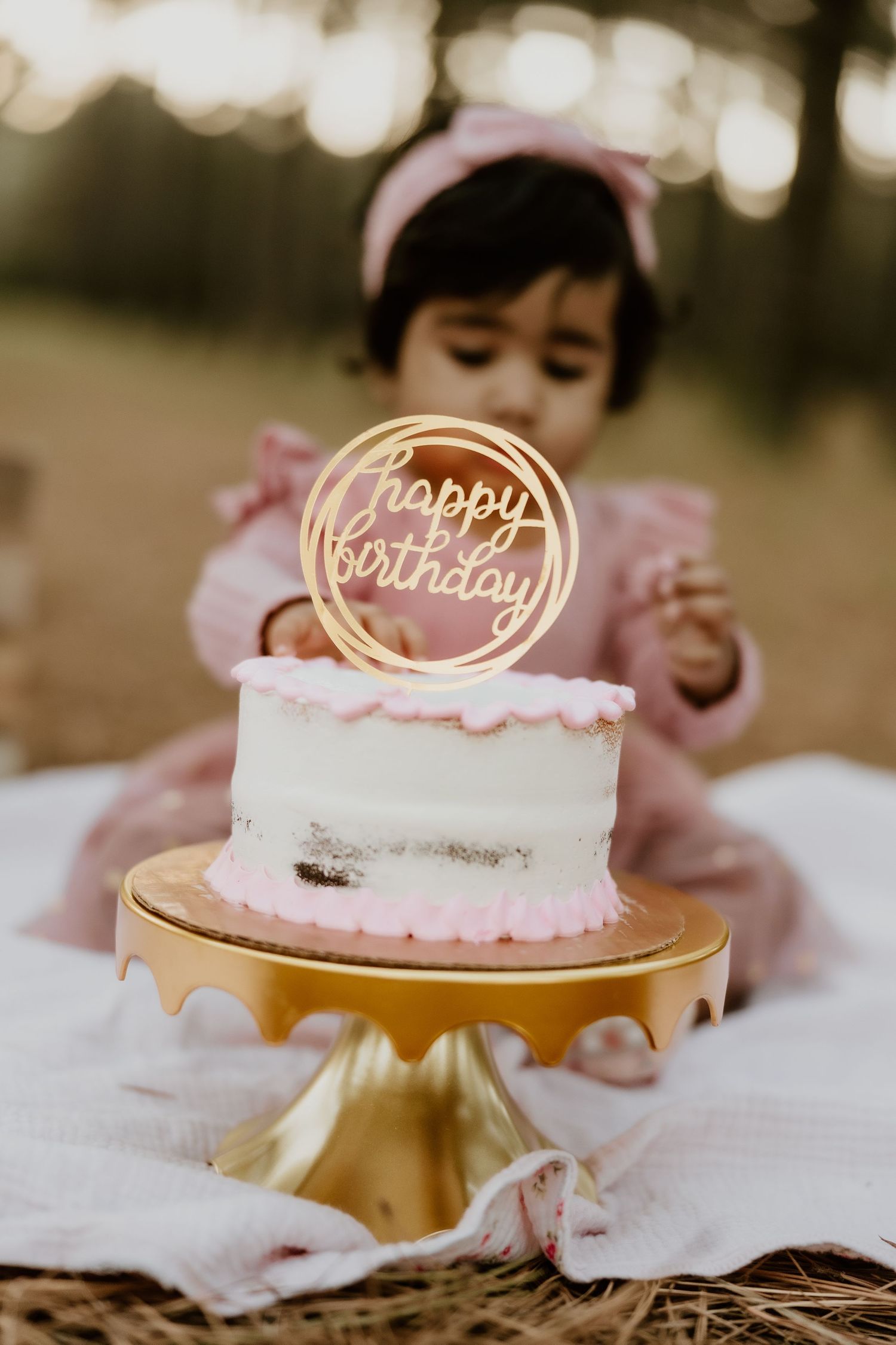 White birthday cake with pink frosting and gold 'Happy Birthday' topper sits on metallic gold cake stand.