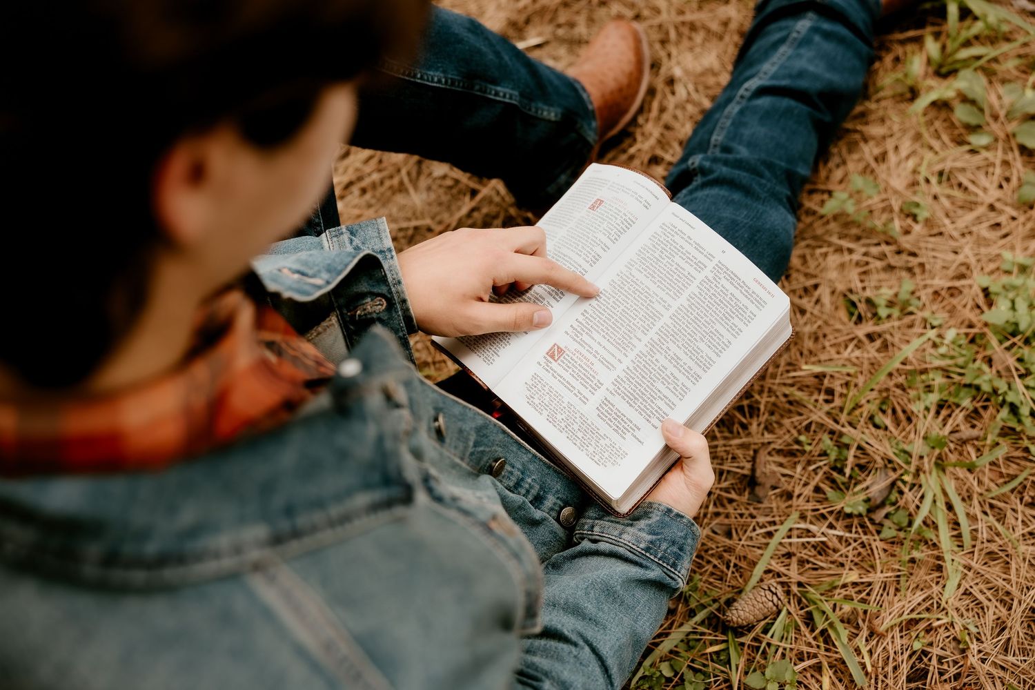 Person sitting outdoors on grass reading a Bible, pointing at a passage while wearing a denim jacket.