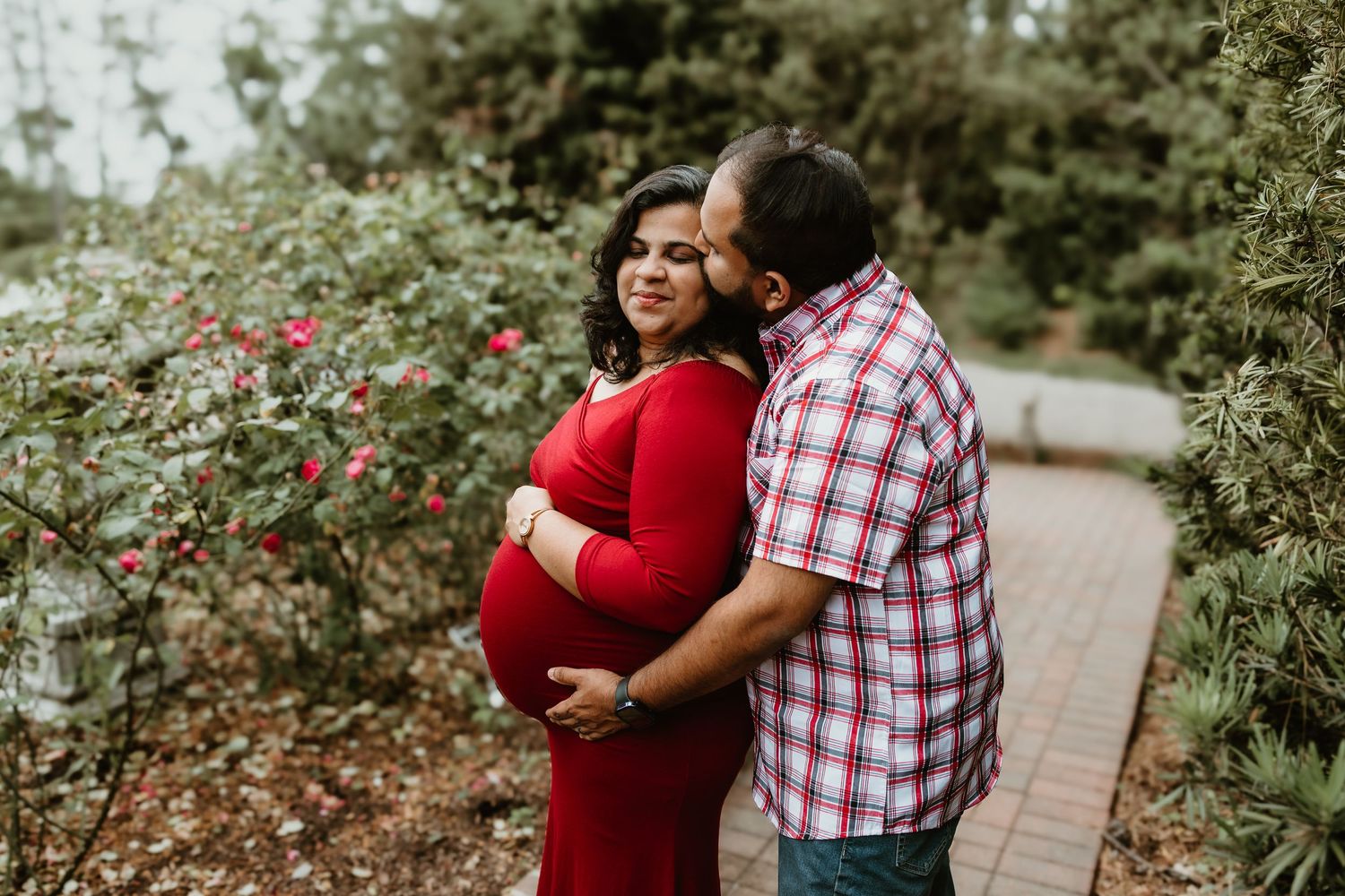 Pregnant woman in red dress embraced by partner in rose garden during outdoor maternity photo session.