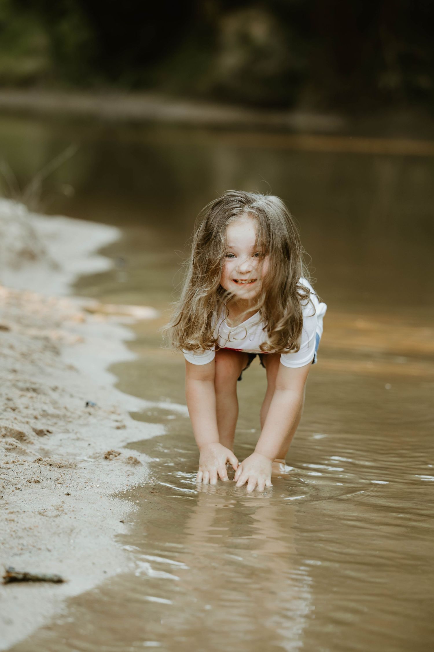 Child in white dress bends down to explore the muddy creek water.