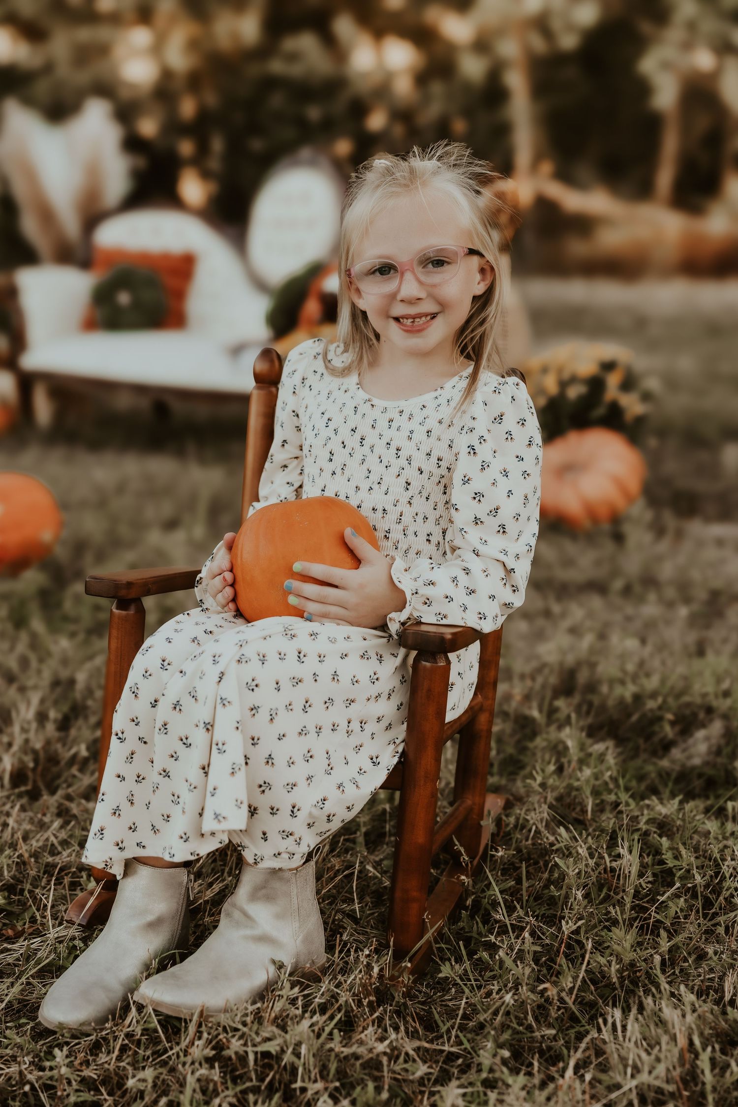 A child in a patterned dress sits in a wooden chair holding an orange pumpkin in an outdoor fall setting.