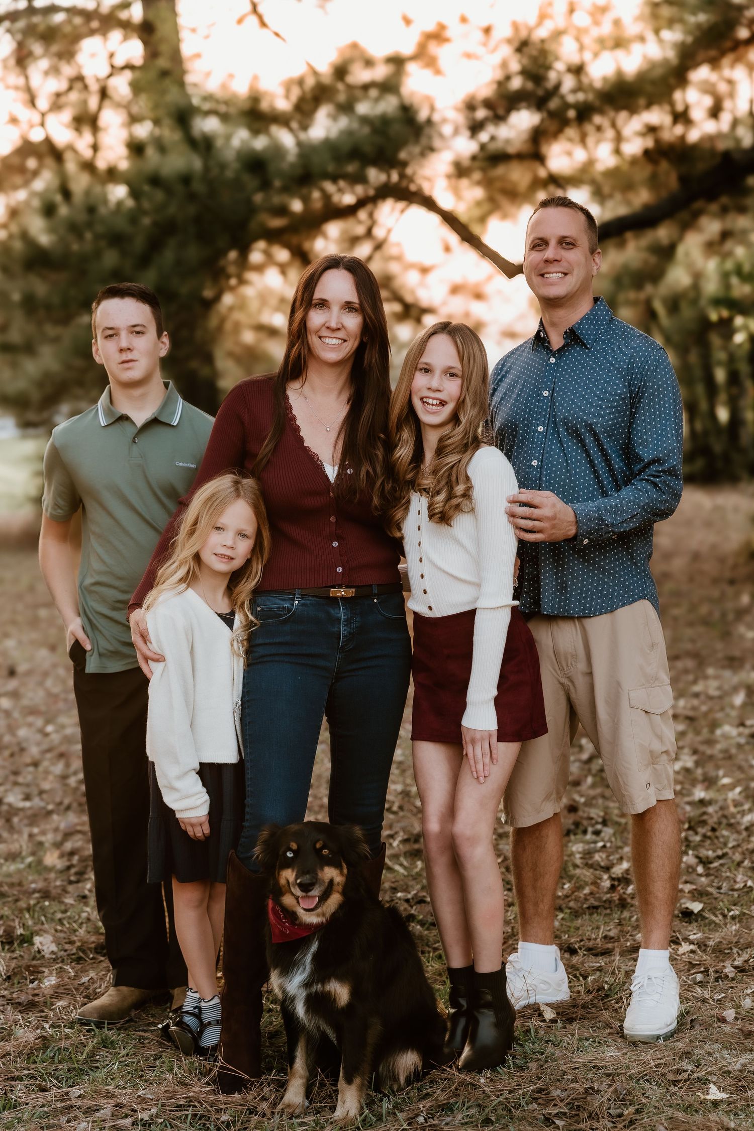 Family portrait in a forest setting at sunset with a black and brown dog standing in front of them.